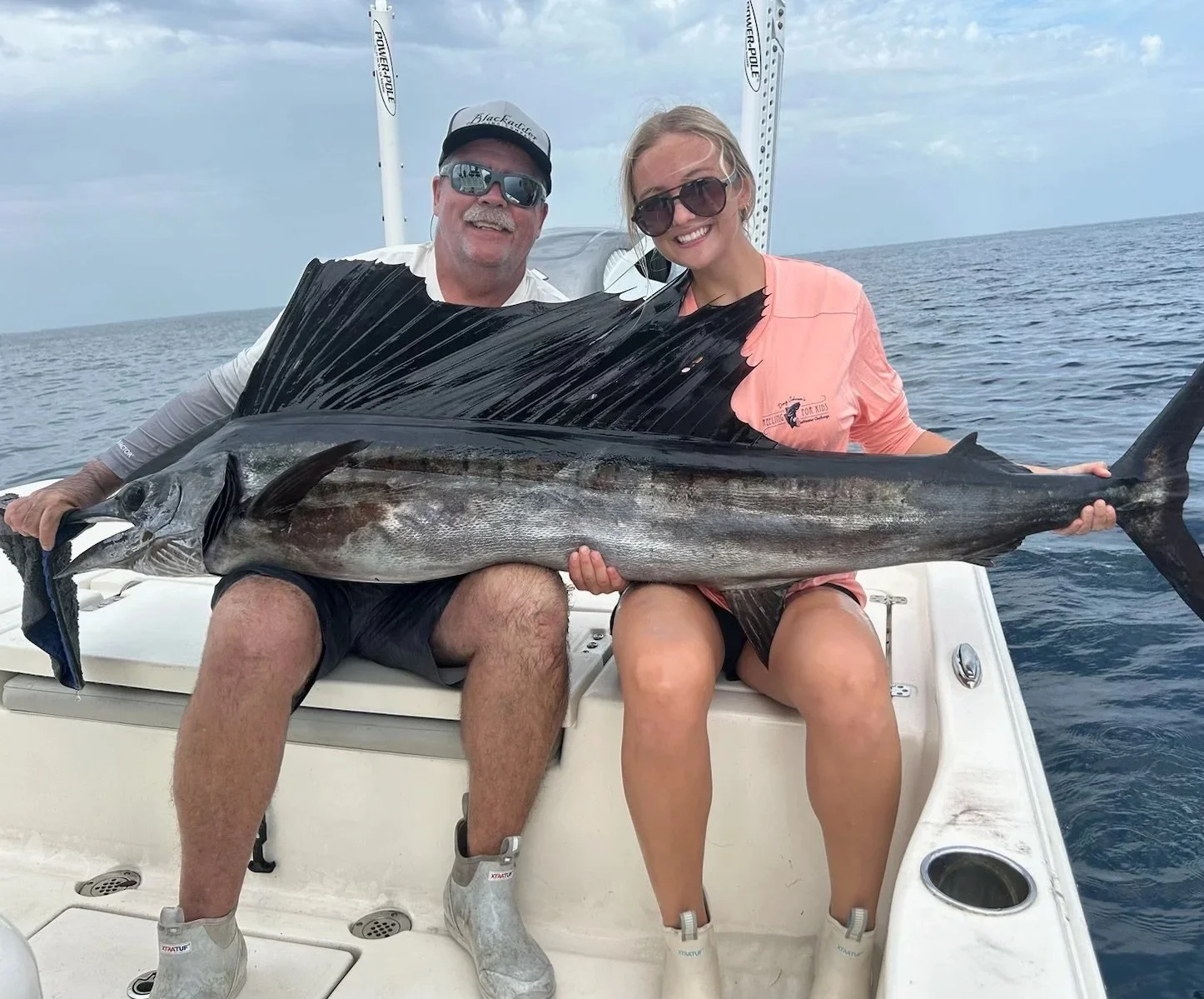 Two people sitting on a boat holding a large fish, with the ocean in the background. The man on the left is wearing sunglasses, a cap, and a long-sleeved shirt. The woman on the right is wearing sunglasses and a coral-colored shirt. They are smiling and appear to be celebrating a fishing success.