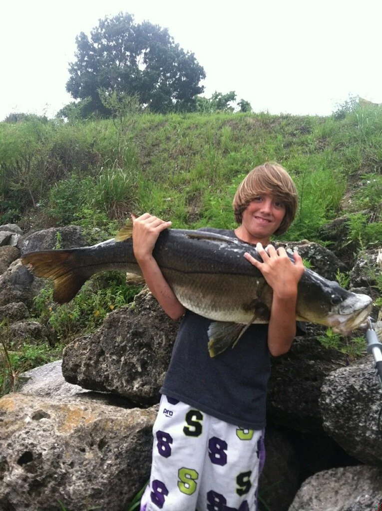 Young boy holding a large fish outdoors, smiling, standing on rocks near greenery and a hillside.