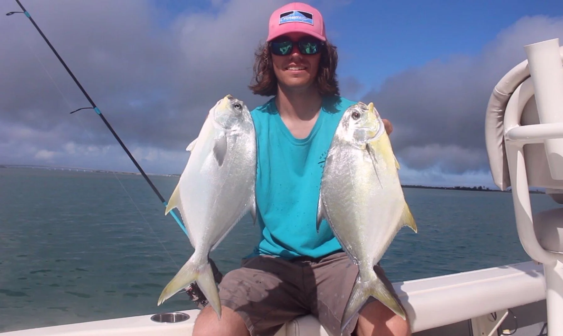 A man with long hair, sunglasses, and a pink cap sitting on a boat, smiling and holding two large fish, one in each hand, with an ocean and cloudy sky background.