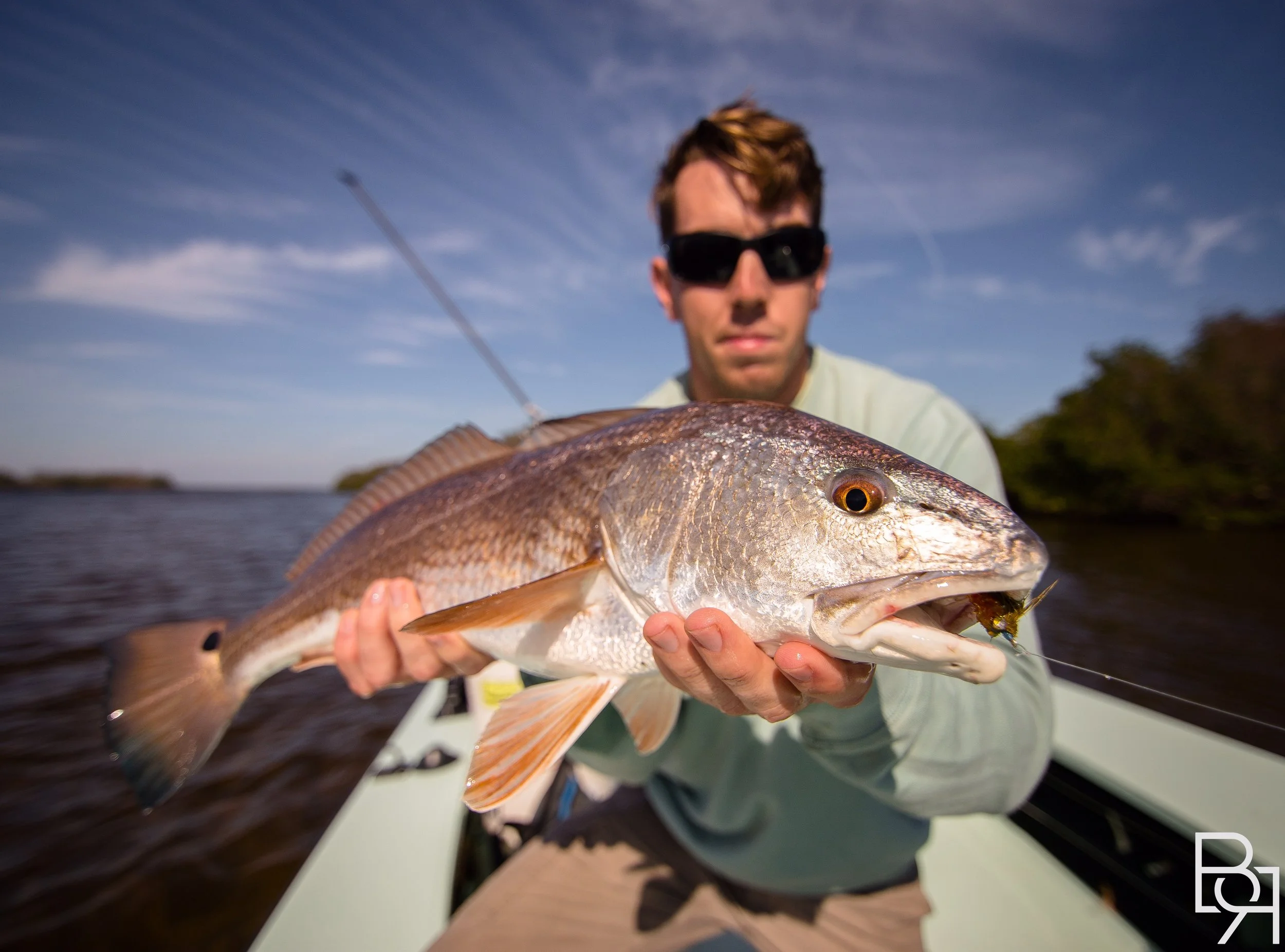 A man with sunglasses holding a large fish on a boat with water and trees in the background.