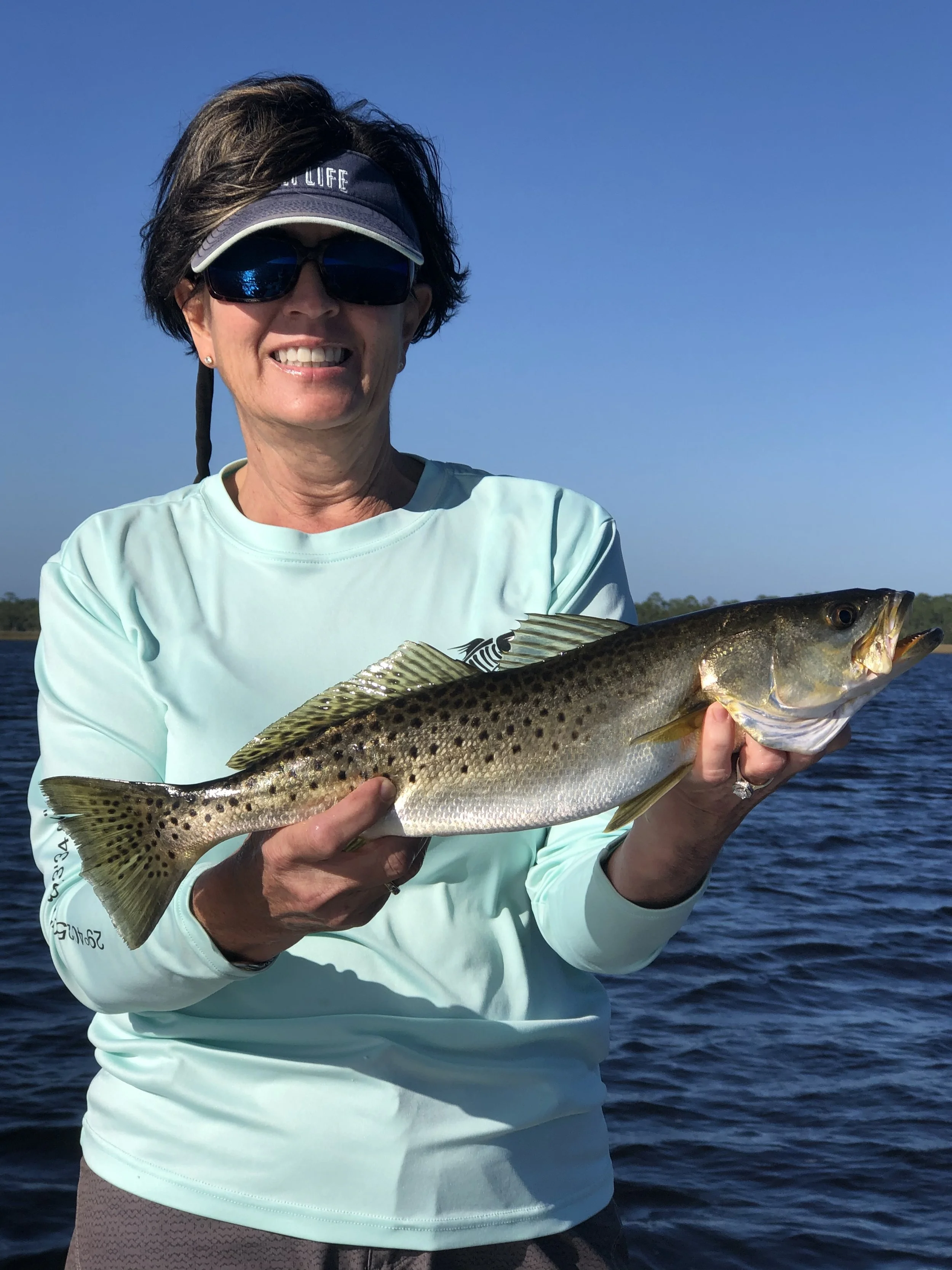 Woman wearing sunglasses and a visor cap holding a large fish over water during daytime.