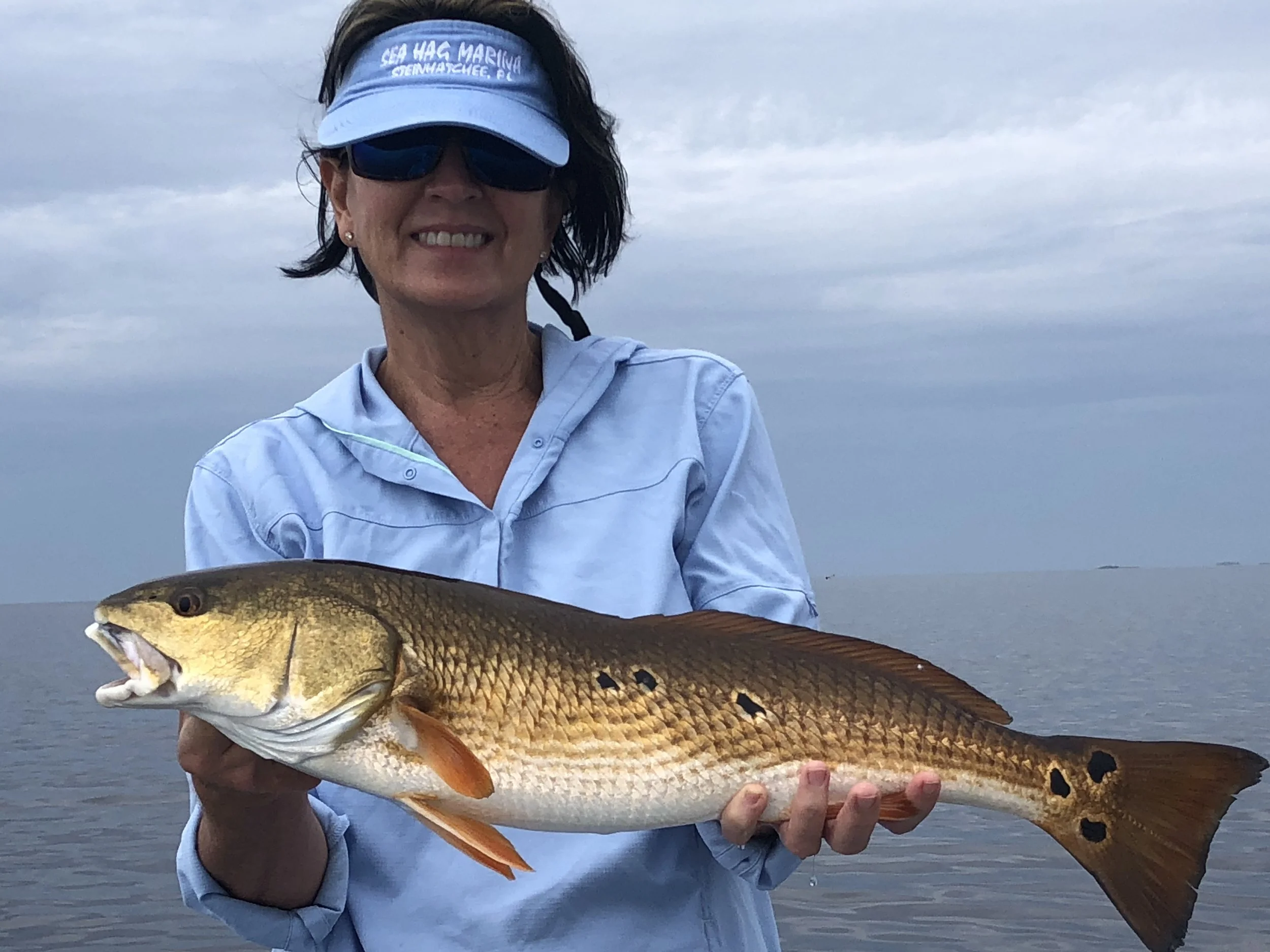 Woman wearing sunglasses and a visor holding a large fish with golden scales and black spots against a cloudy sky and water background.