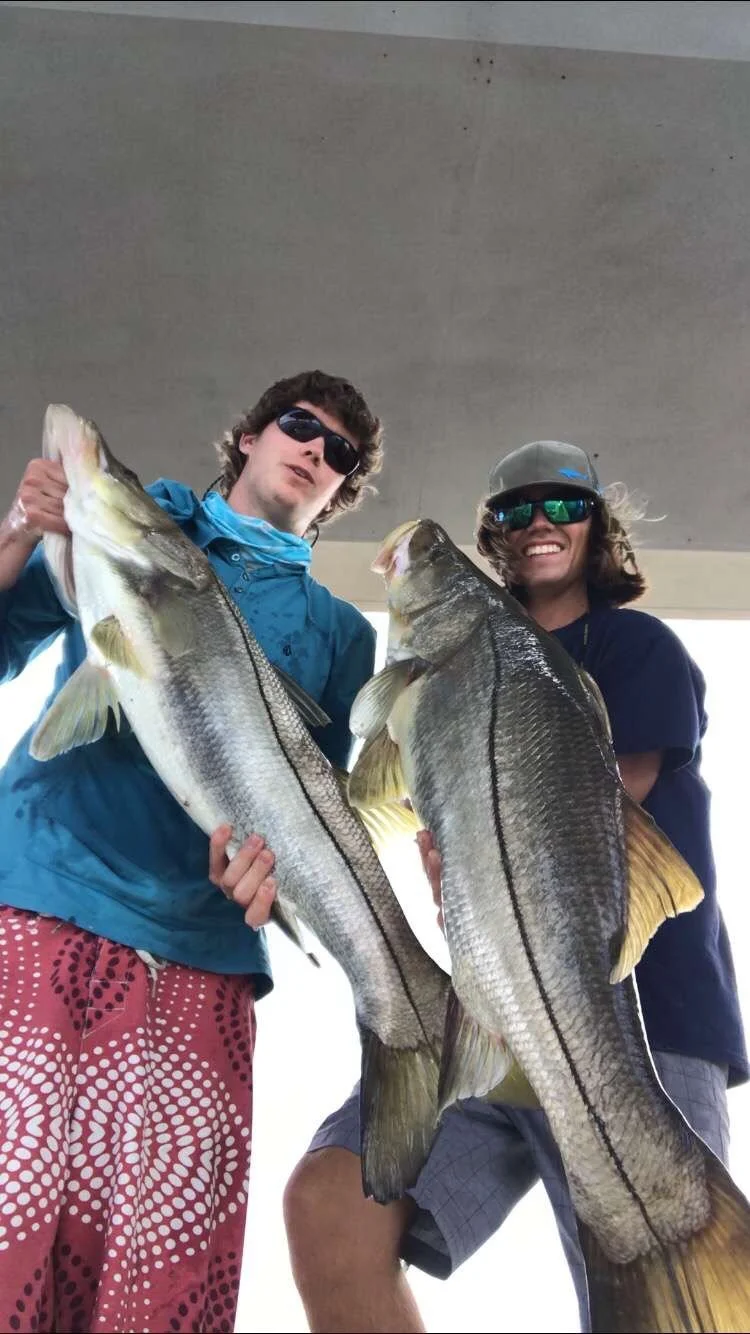 Two young men holding large fish, smiling, wearing sunglasses, one in a blue jacket and red patterned shorts, the other in a dark shirt and gray shorts, under an overpass.