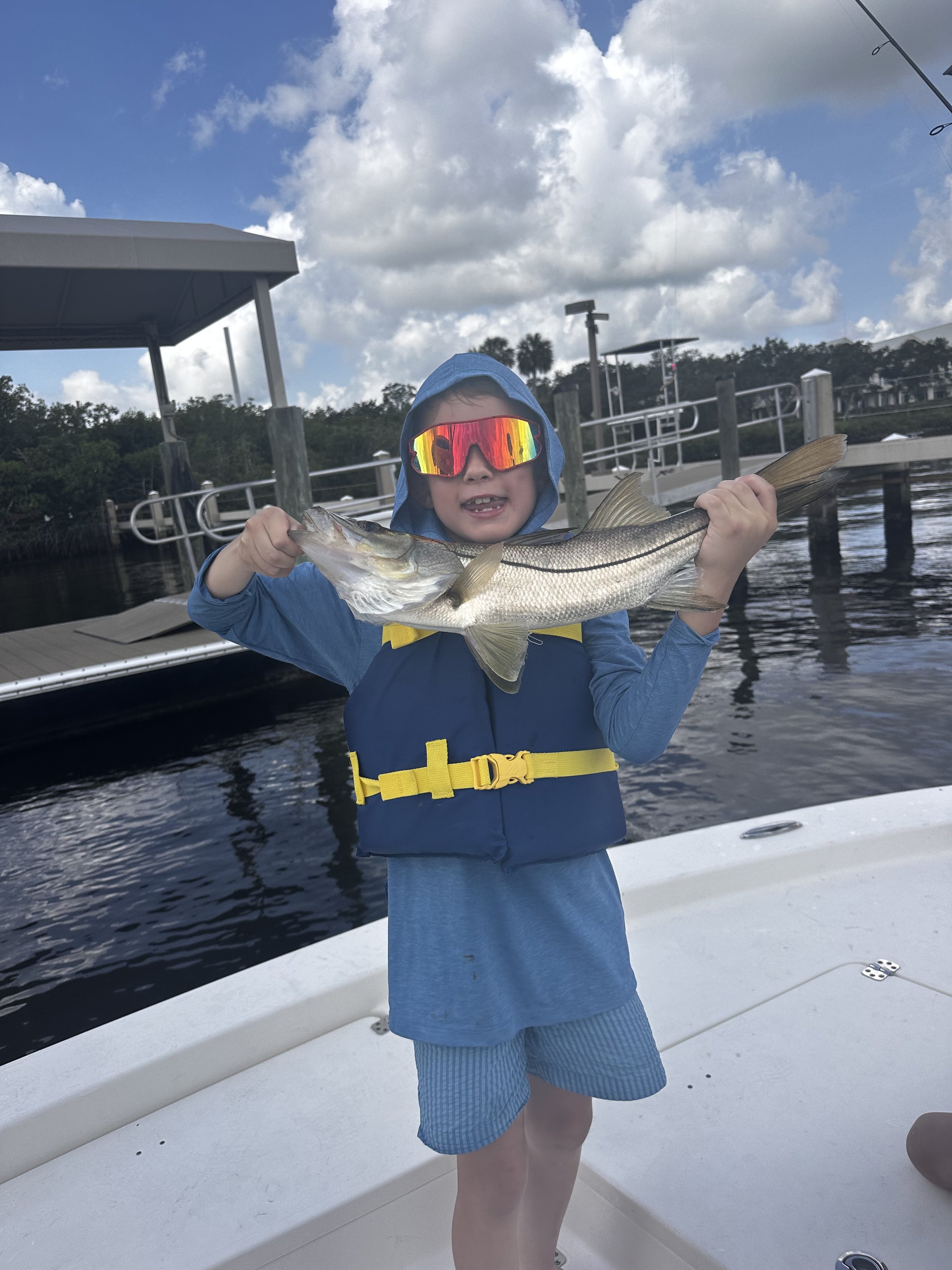 A young boy wearing a blue hooded jacket, striped shorts, and mirrored sunglasses is holding a large fish aboard a boat. The background features a marina with docks, boats, and a partly cloudy sky.