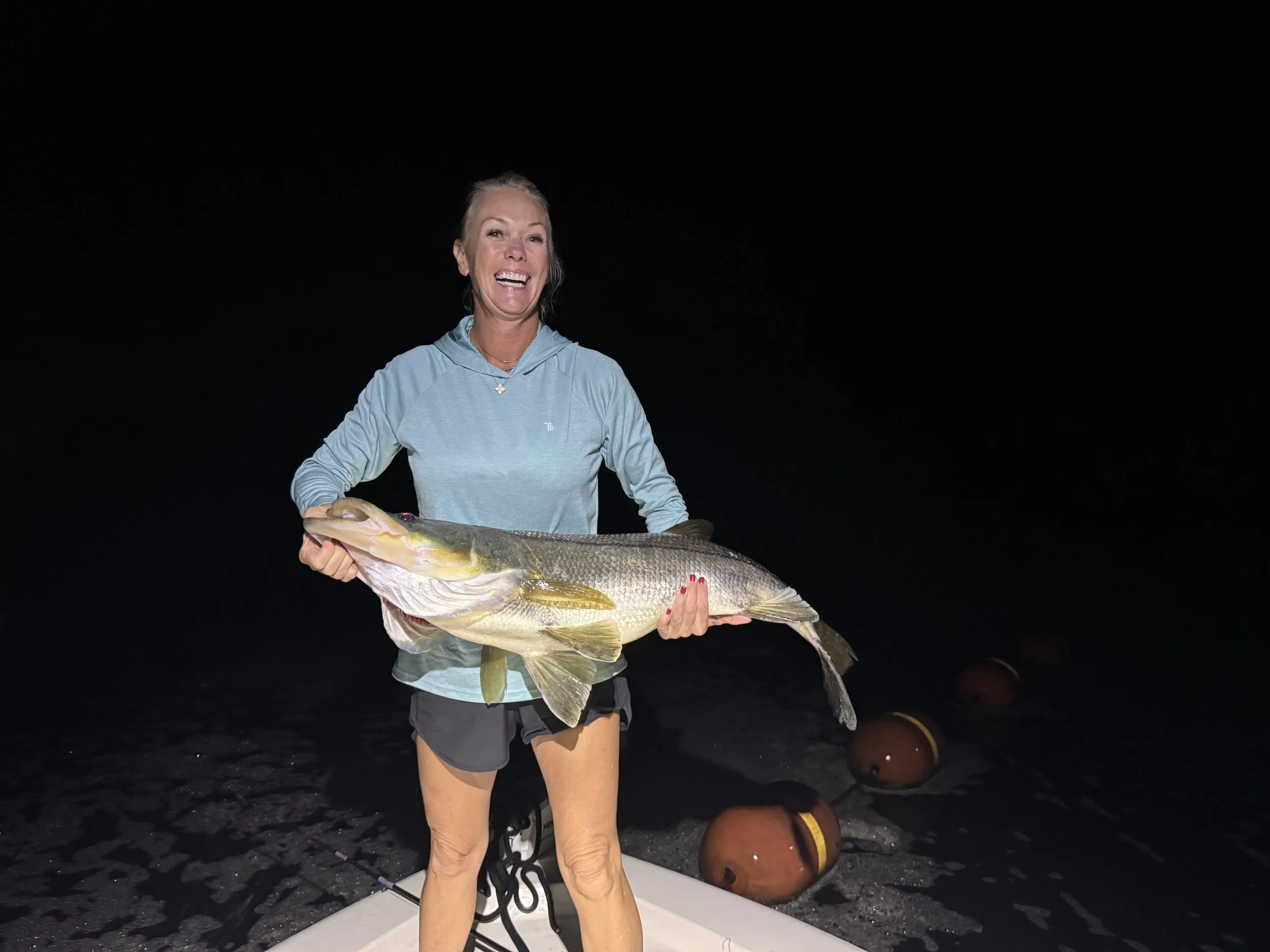 A woman in a light blue hoodie and black shorts is holding a large fish, likely a cod, during nighttime. She is smiling and standing on a boat or dock with orange buoys or floaters and dark water in the background.