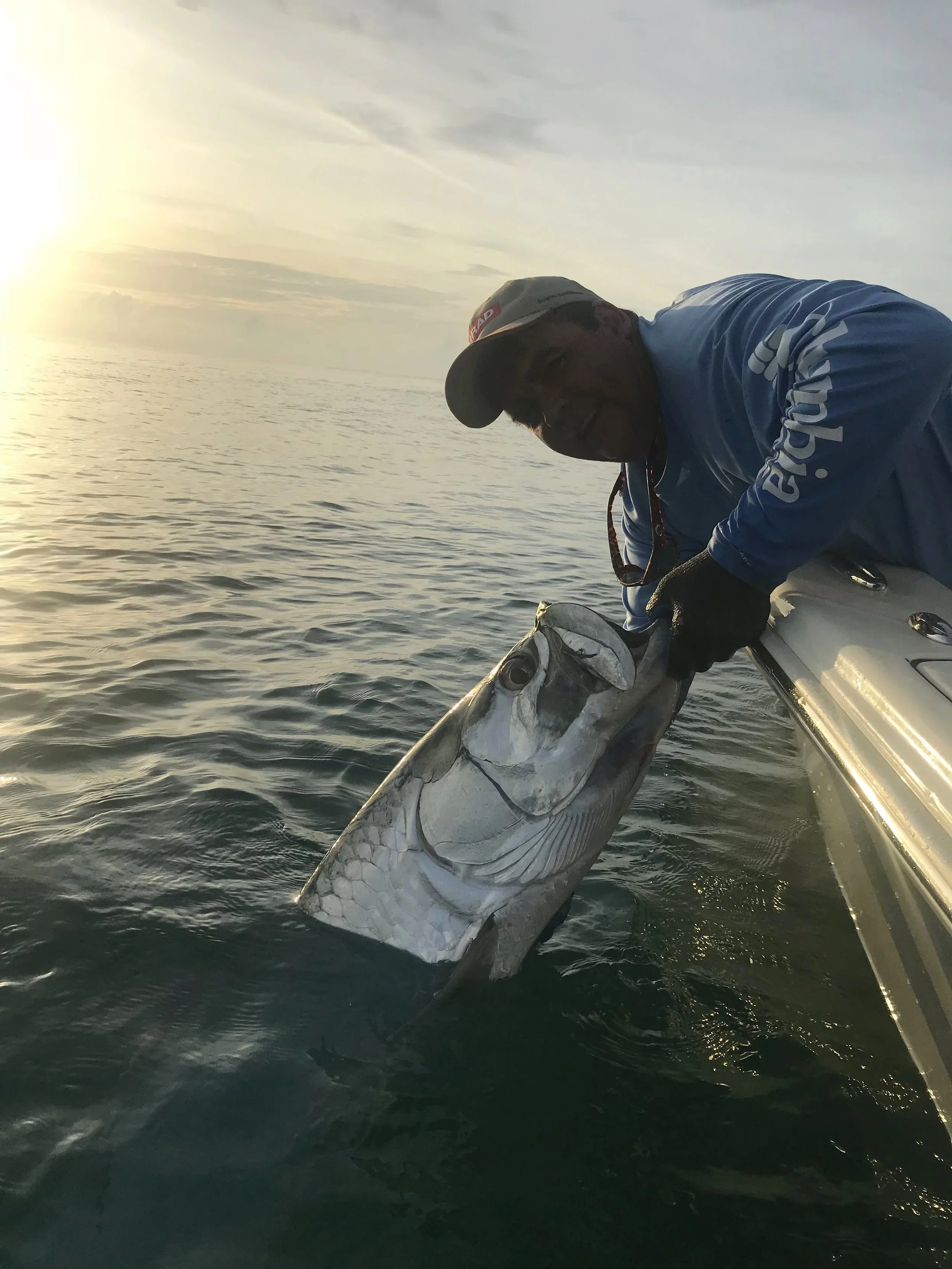 A man in a blue jacket and cap holding a large fish on a boat at sunset.