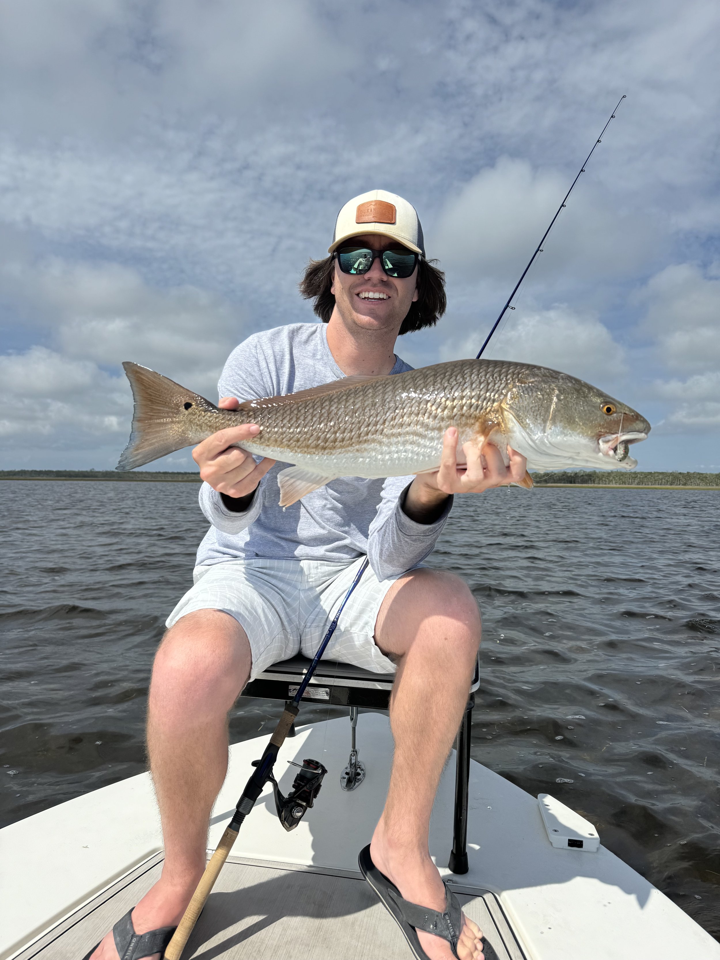 A man smiling on a boat holding a large fish he caught while fishing. He is wearing sunglasses, a baseball cap, and casual clothes, with water in the background.