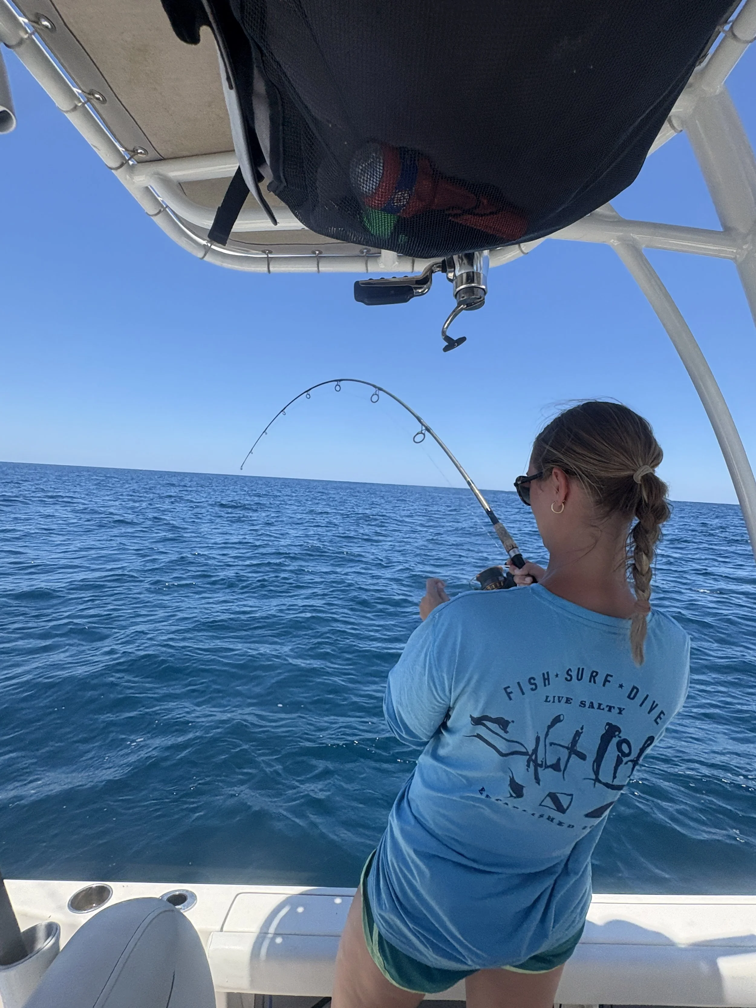 A woman with braided hair wearing sunglasses and a blue T-shirt fishing off a boat on the ocean with a clear blue sky.