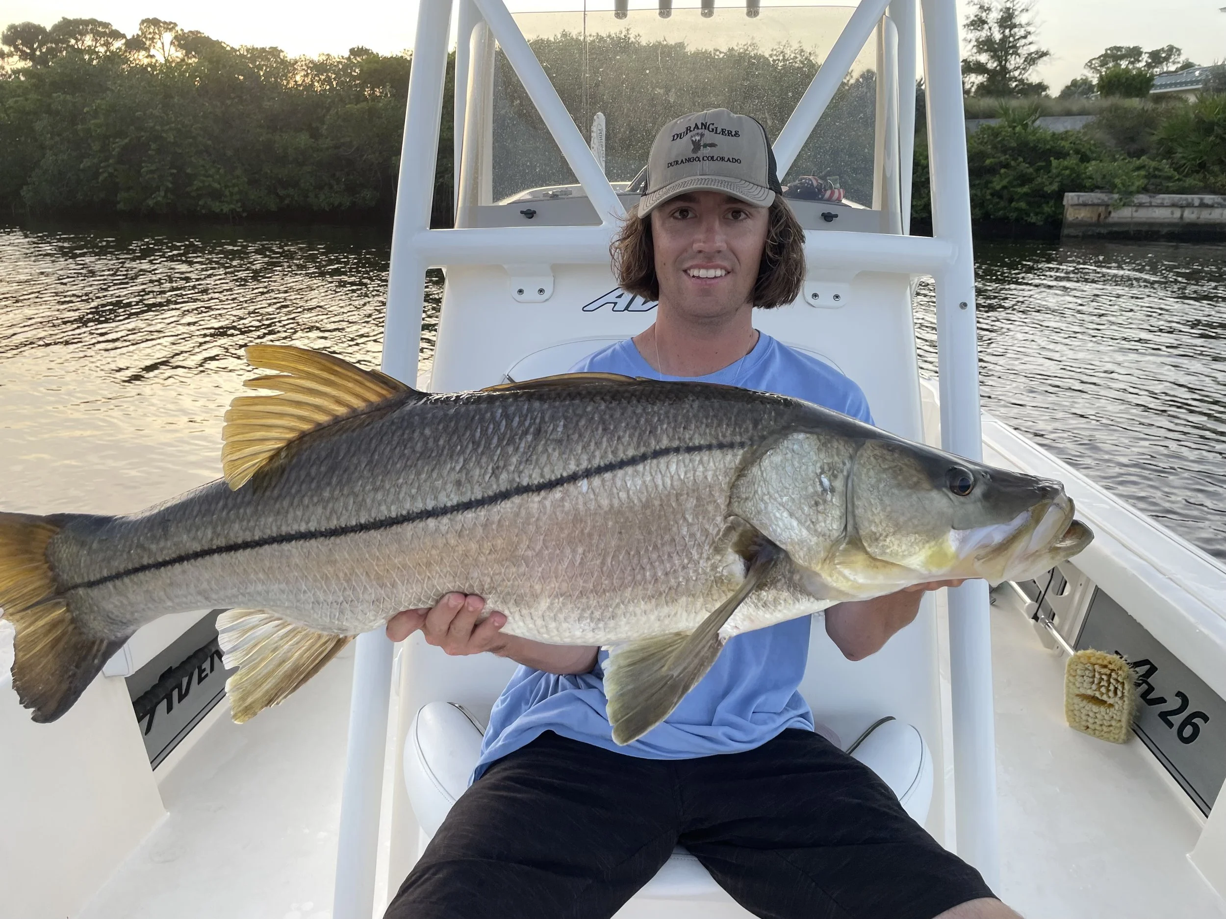 A man with long hair wearing a cap and blue shirt, sitting in a boat, holding a large fish he caught, with water and greenery in the background during sunset.