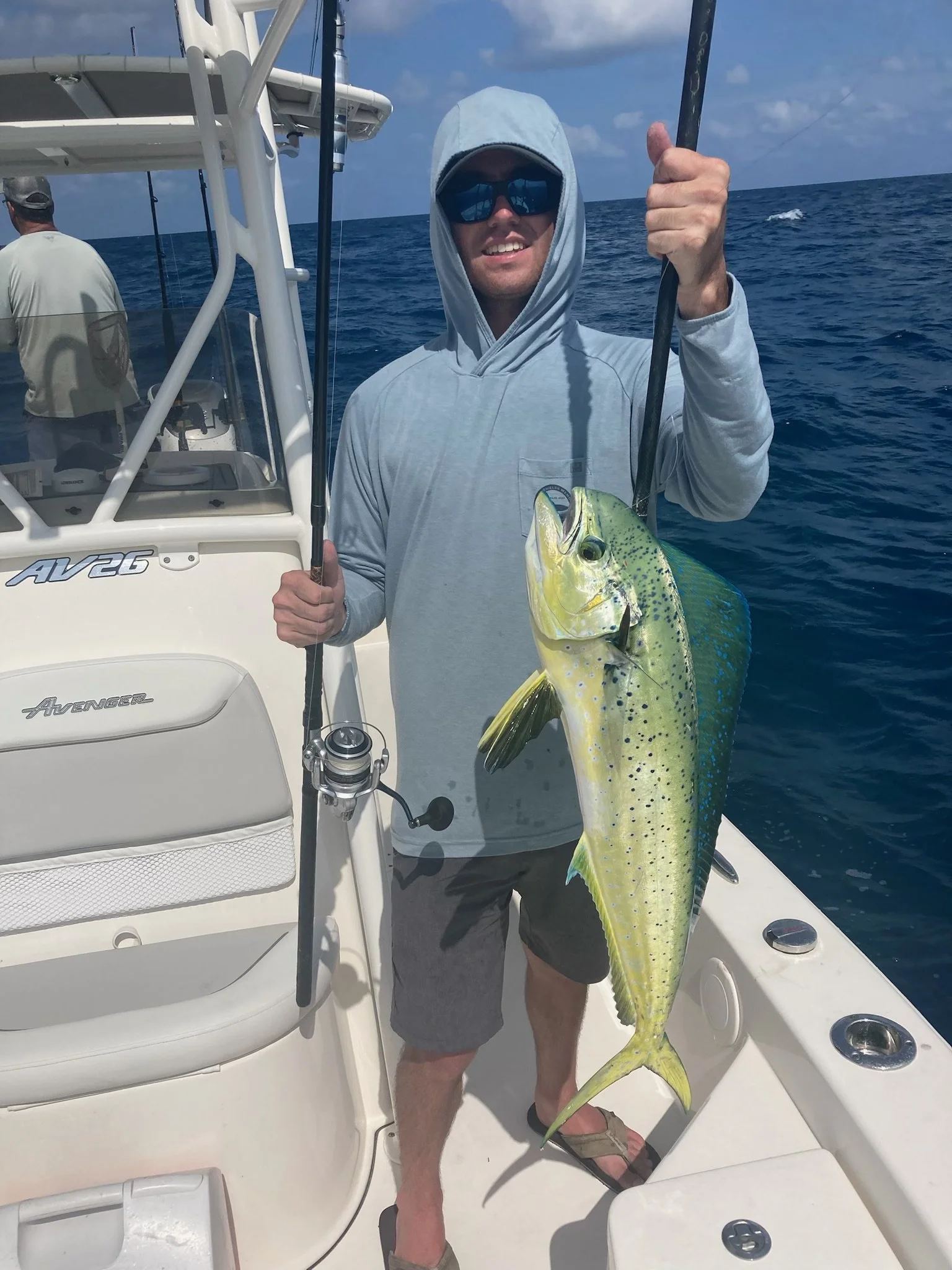 A man on a boat holding a large fish he caught, with another person in the background and the open ocean around them.