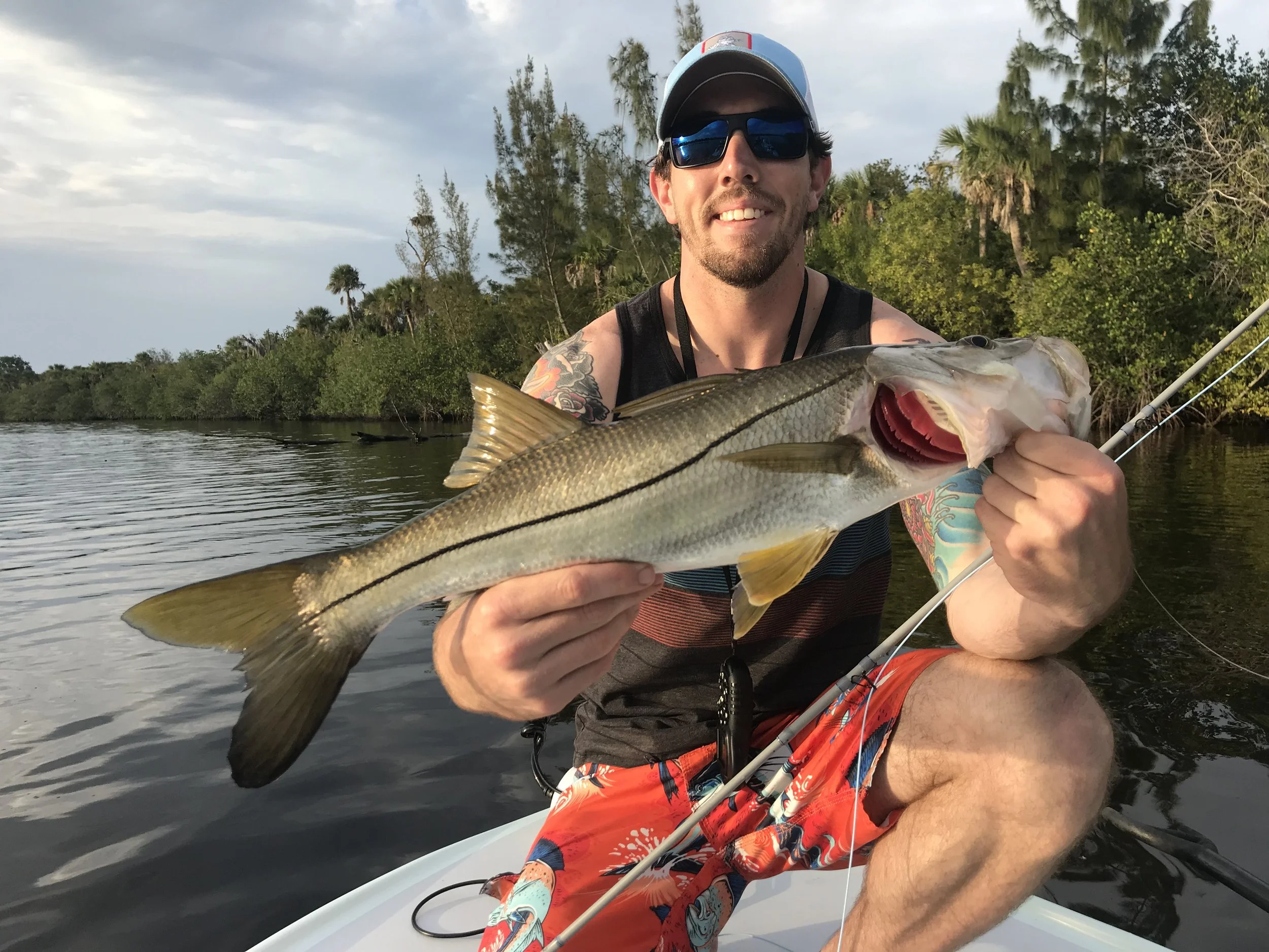 A man smiling while holding a fish he caught on a boat with lush greenery and trees in the background.