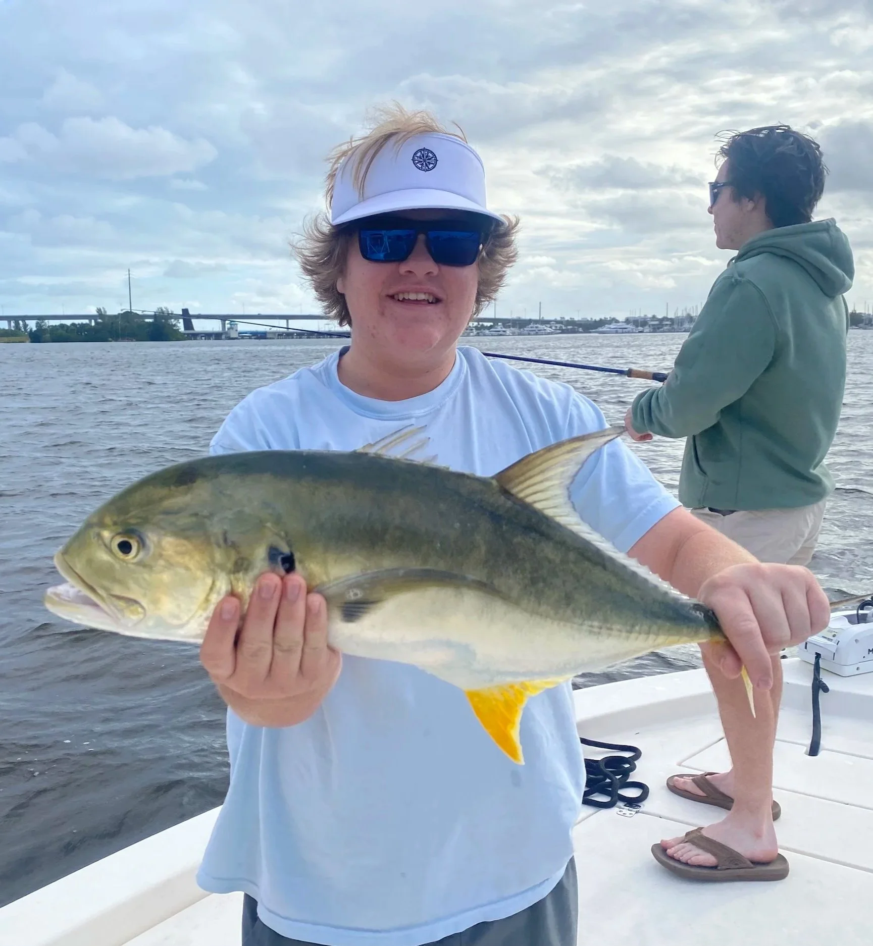 A young man with blonde hair wearing sunglasses and a white visor cap holding a fish on a boat. In the background, another person is fishing, and there are boats and a bridge visible in the distance with a cloudy sky.