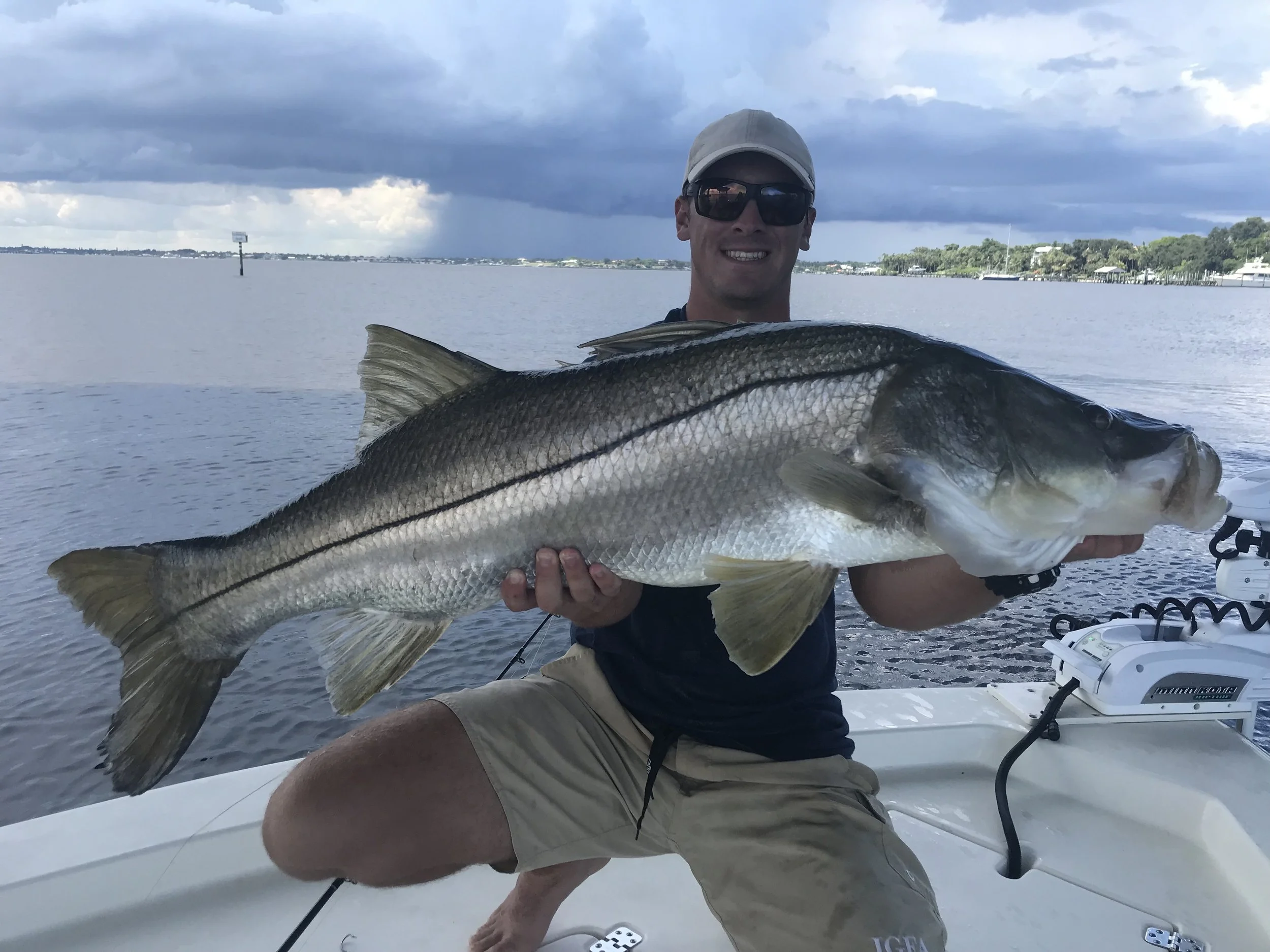 A man sitting on a boat holding a large fish in front of him, with water and cloudy sky in the background.