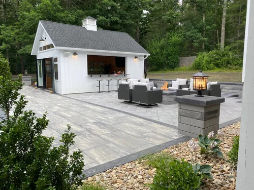 Outdoor patio with seating area and a small white building, surrounded by greenery and rocks,