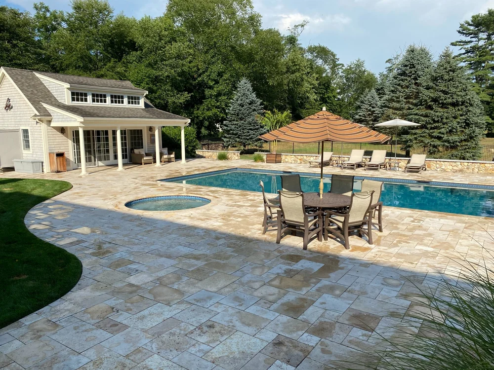 A backyard with a swimming pool and hot tub, featuring patio furniture, umbrellas, and a pool house, surrounded by trees and greenery.