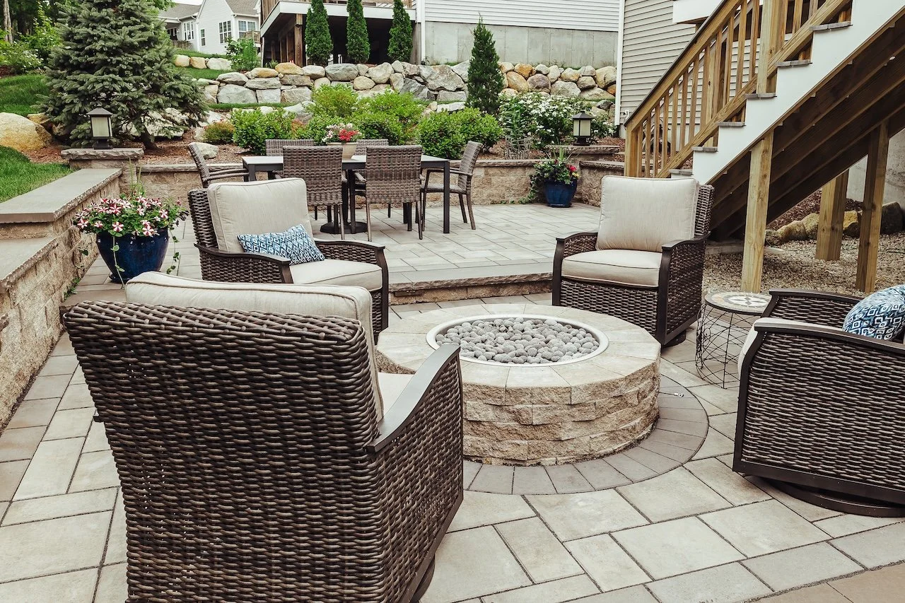 A backyard patio area featuring outdoor seating with wicker chairs and cushioned armchairs surrounding a circular stone fire pit filled with rocks. In the background, there is a dining table with wicker chairs on a raised stone platform, with decorative plants and a rock garden behind. A staircase with wooden railing leads up to the house. There are potted plants with flowers, and the yard is landscaped with trees, shrubs, and stone walls.