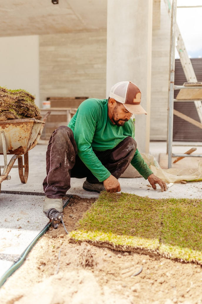 A man laying sod on a construction site with scaffolding and building materials in the background.
