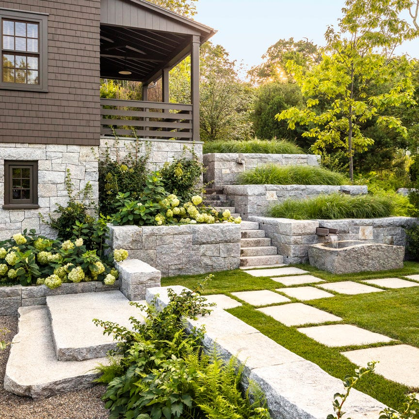 A landscaped backyard with stone steps, lush green grass, and trees, adjacent to a house with a small balcony.
