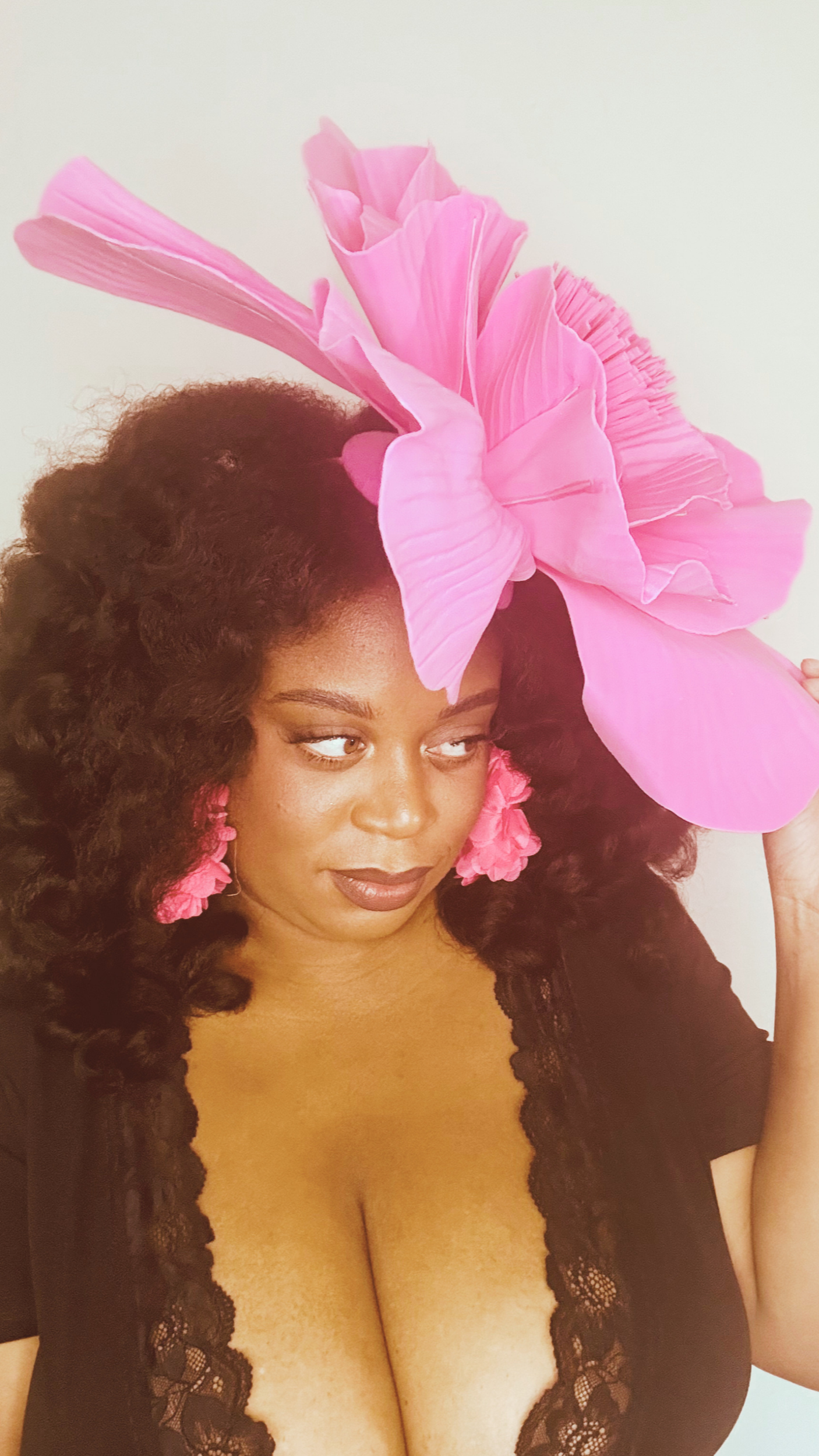 A woman with dark curly hair and makeup, wearing black lace clothing and pink floral earrings, holding a large pink flower above her head.