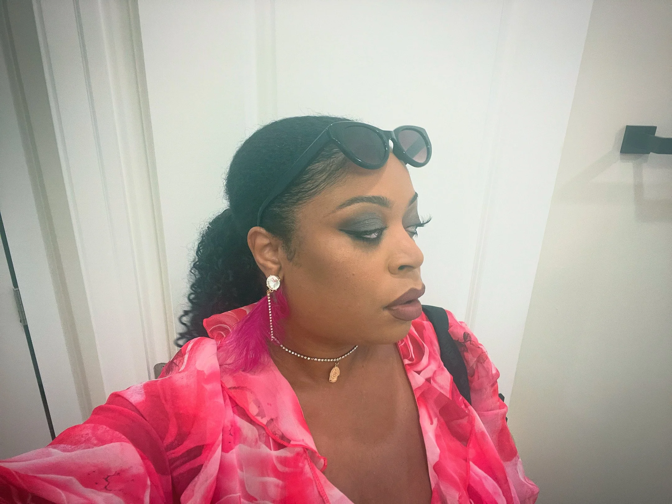 A woman with dark hair styled in loose curls, wearing sunglasses on her head, earrings, a necklace, and a pink patterned top, standing in front of a white wall.