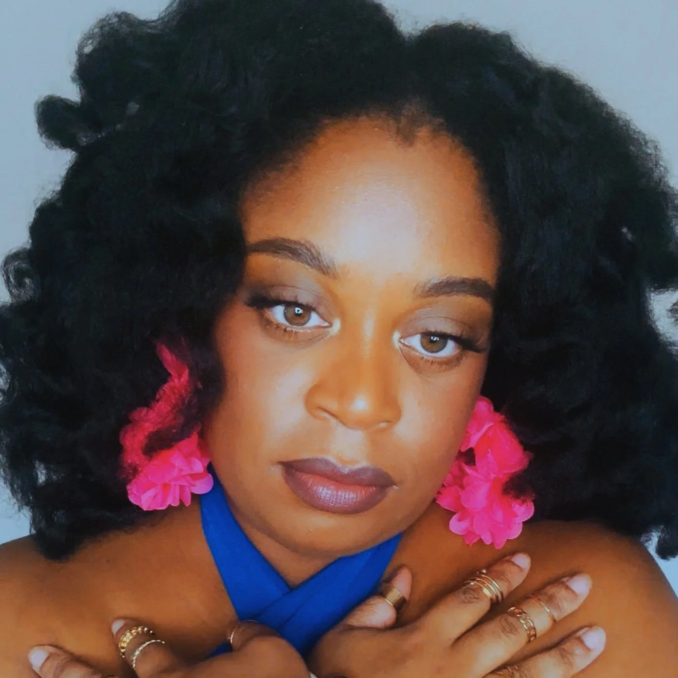 A woman with curly black hair, brown eyes, and dark lipstick. She is wearing pink flower earrings, multiple rings, and a blue halter top, posing with her hands crossed over her chest against a plain background.
