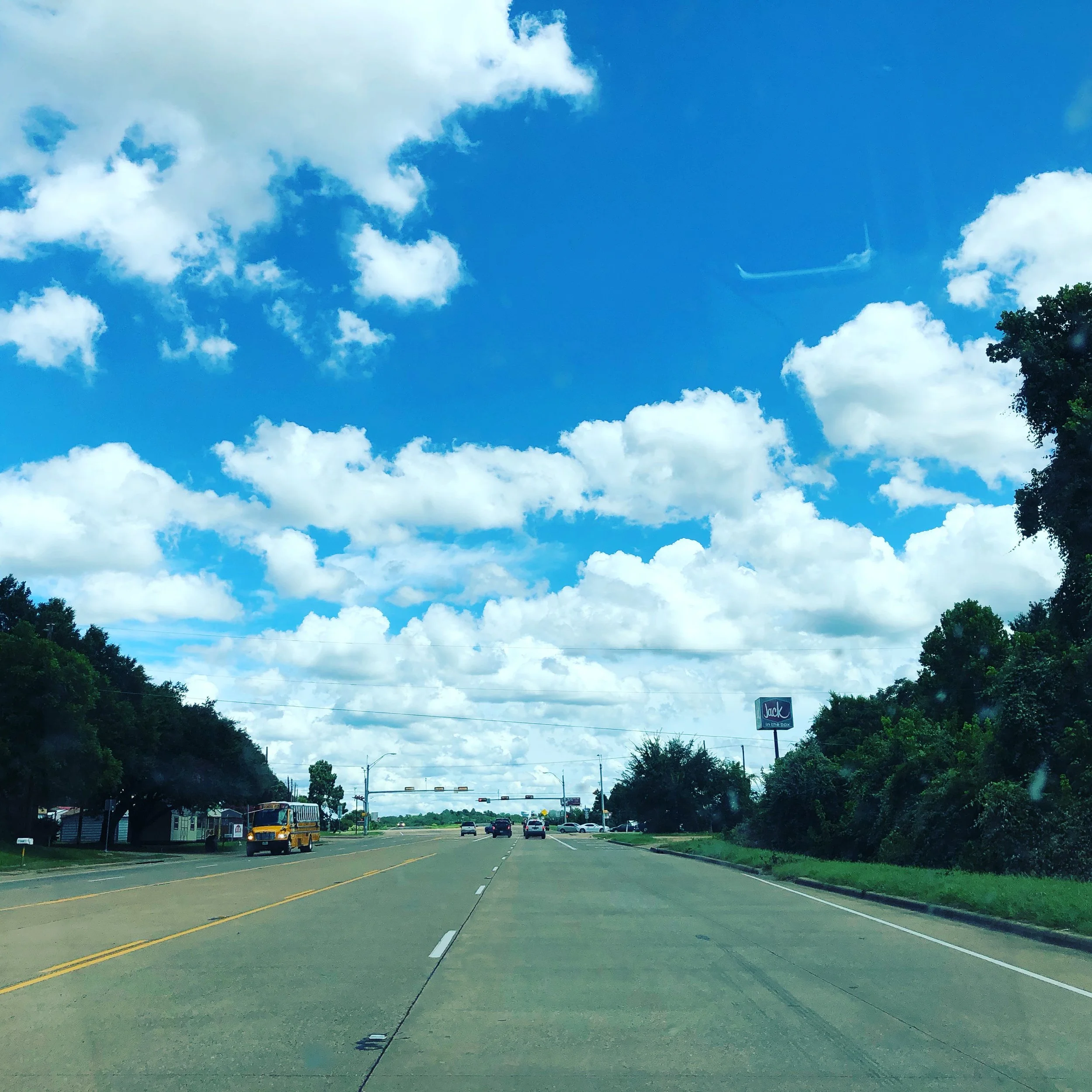 A wide view of a suburban street with a traffic light and cars parked on the side. The sky is mostly blue with scattered white clouds, and trees line the street. A billboard with the word 'Jack' is on the right side of the image.