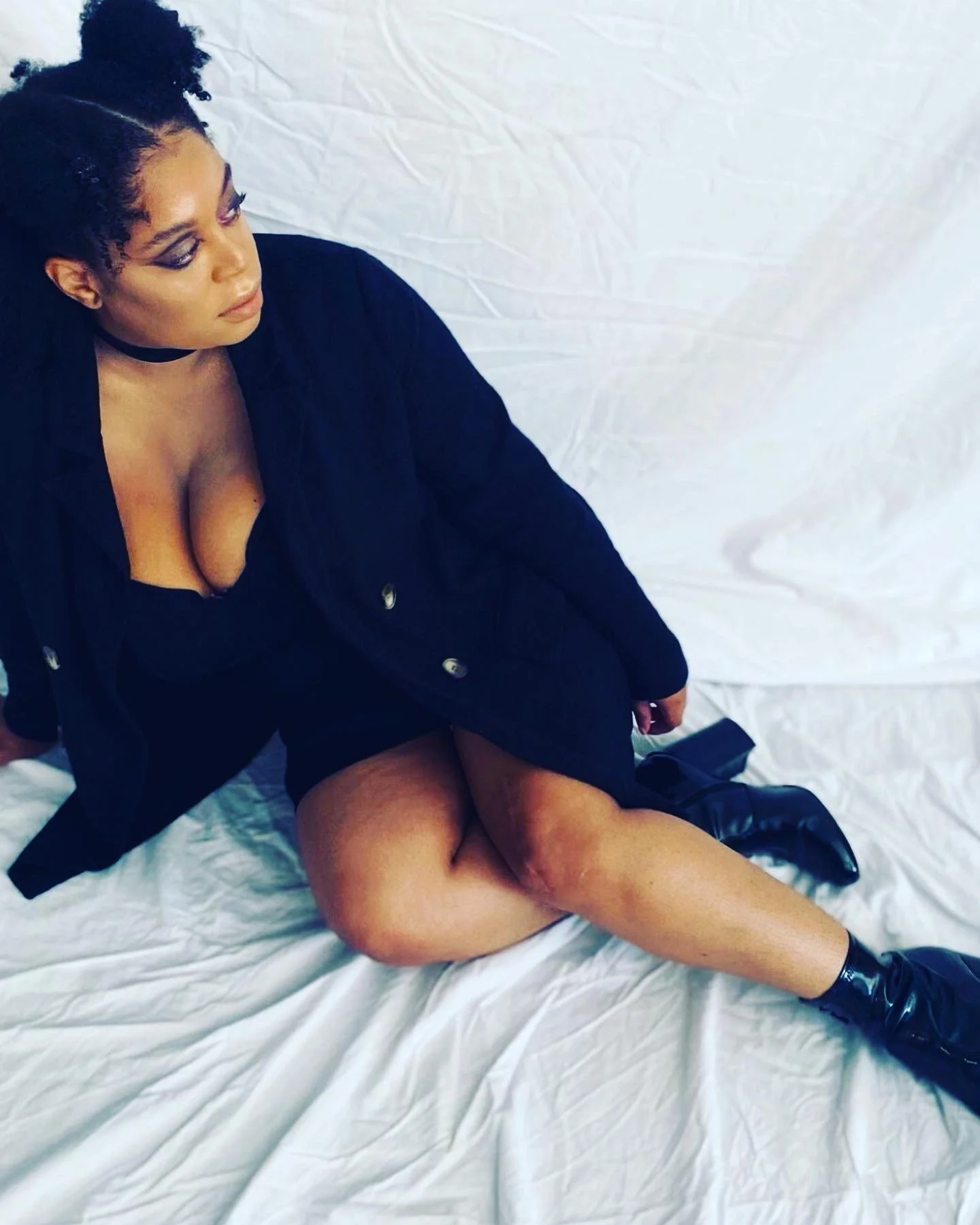 Woman with dark hair styled in twists and a bun, wearing makeup, a black choker, black jacket, black dress, black boots, and stockings, sitting on a white crinkled fabric surface, with her legs spread and looking down.