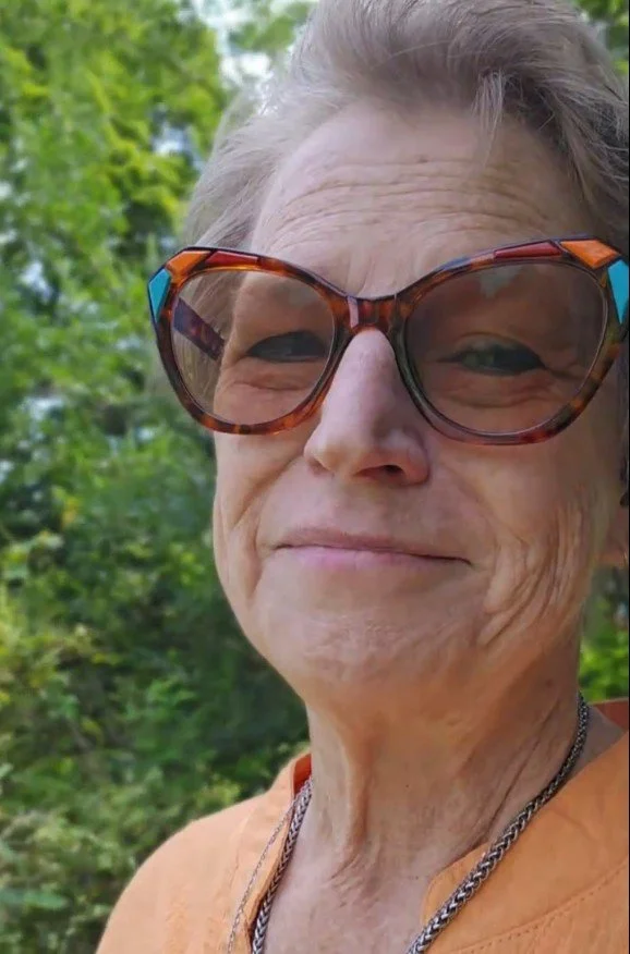 Close-up of an elderly woman outdoors wearing large tortoiseshell sunglasses, smiling slightly, with green foliage in the background.