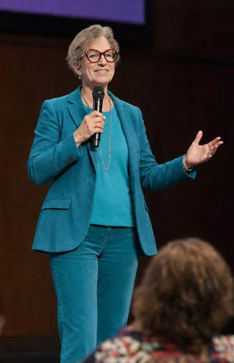 A woman with short hair wearing glasses, a teal blazer, and teal jeans is speaking into a microphone on stage, gesturing with her right hand, with an audience member in the foreground.