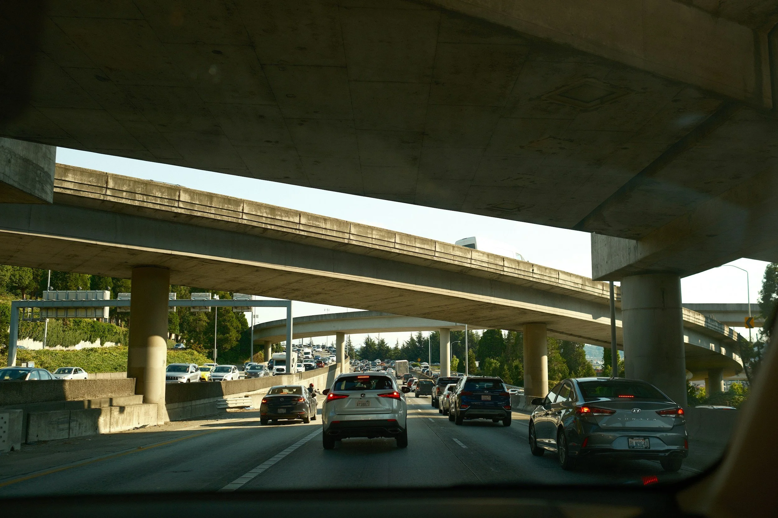 Multiple lanes of traffic with cars traveling underneath a large concrete overpass on a highway, with more cars and greenery visible in the background.