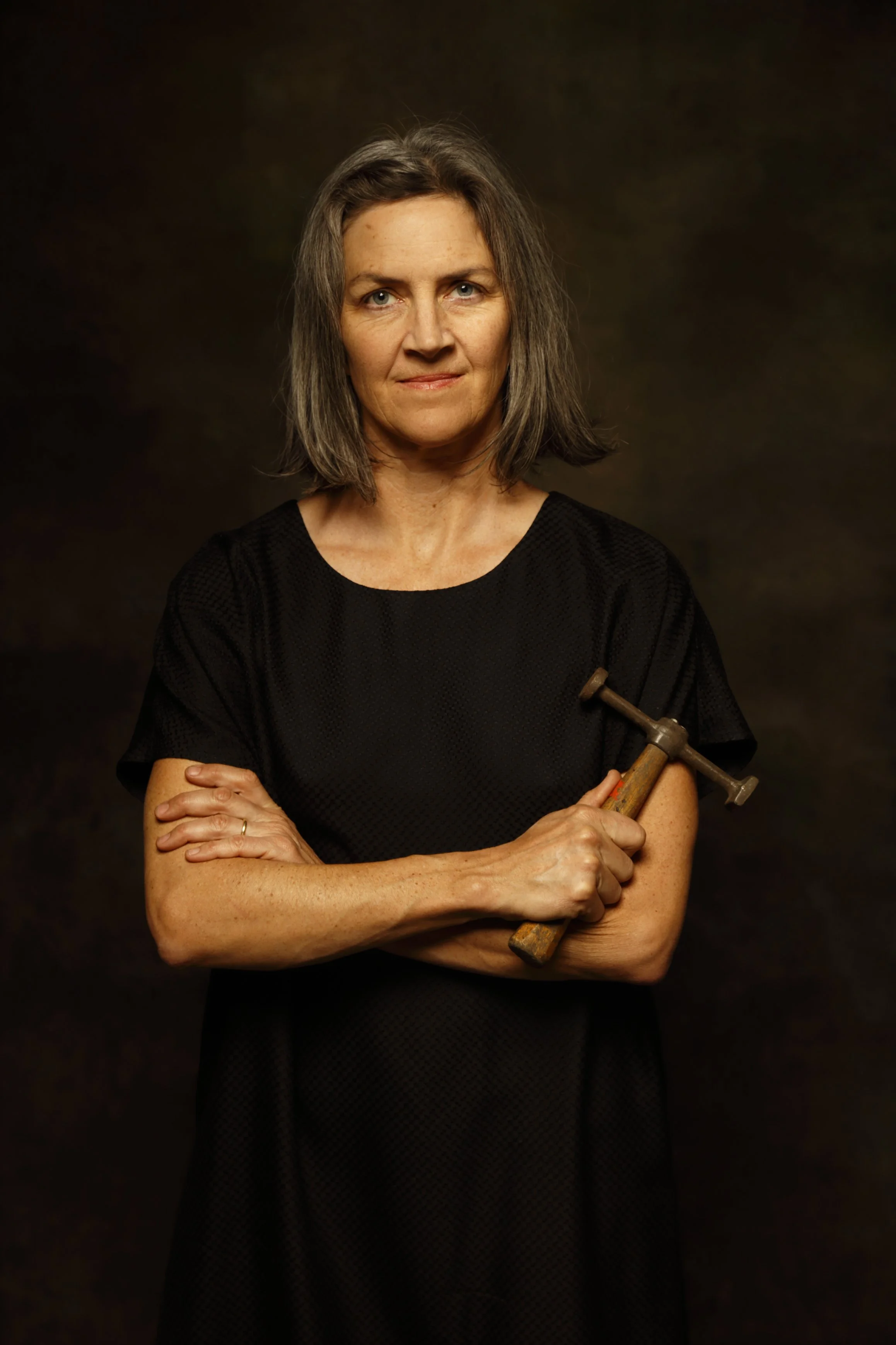 A middle-aged woman with shoulder-length gray hair, wearing a black dress, is standing against a dark background. She is holding a large adjustable wrench with her right hand across her body and has her left arm crossed over her chest.