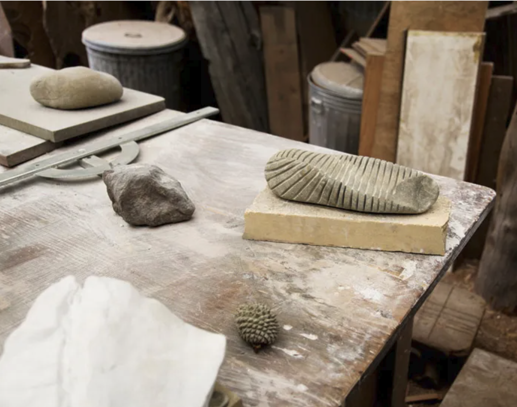 Worktable with stones, clay, and tools for sculpture; background shows workshop with trash can and wood panels.