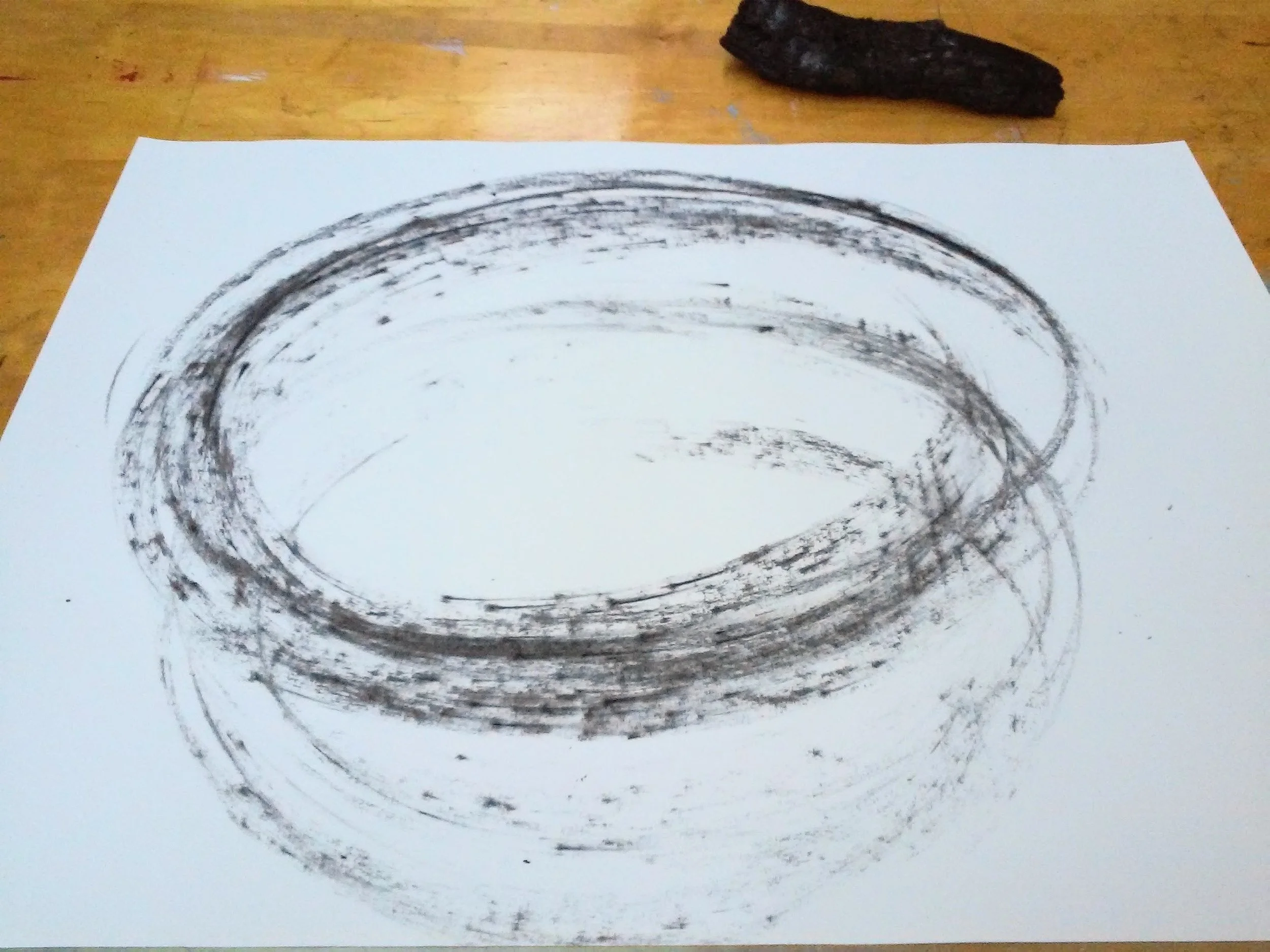 A drawing of circular energies on a white sheet of paper, placed on a wooden surface, drawn with black charcoal from the Vision Fire, Inverness, California.