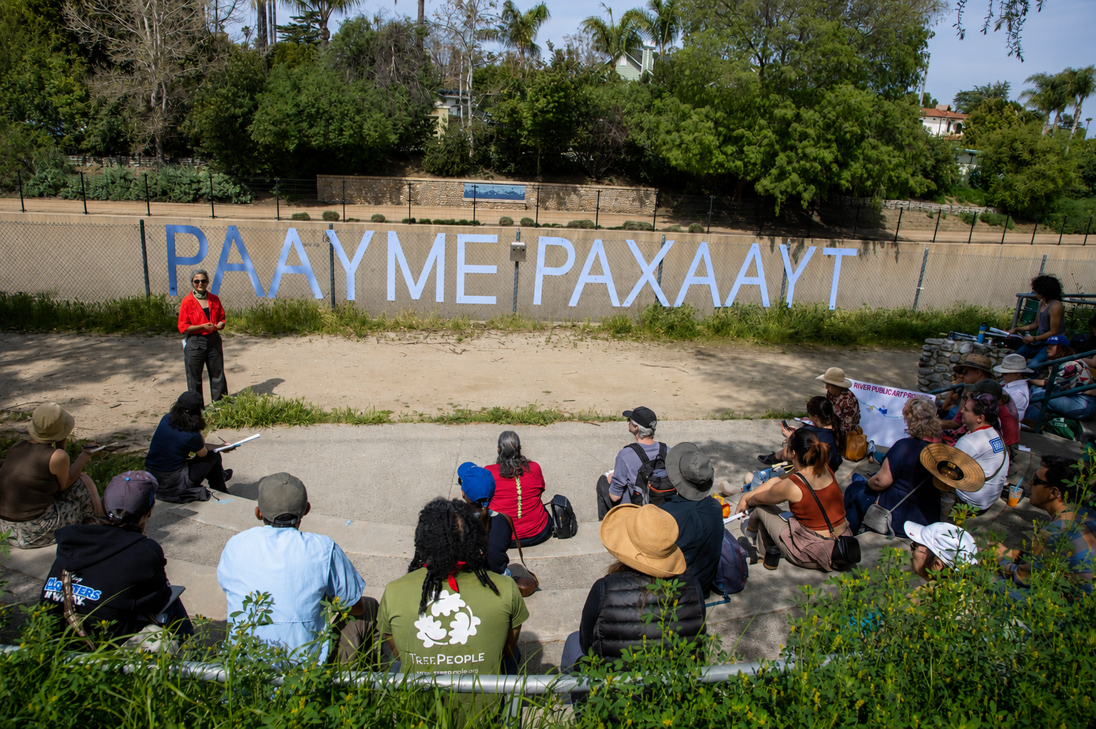 A group of people sitting on steps outdoors, listening to a woman in red speak. Behind her, a fence with large blue letters spelling 'PAY ME PAXAAYT'. Trees and houses are in the background.