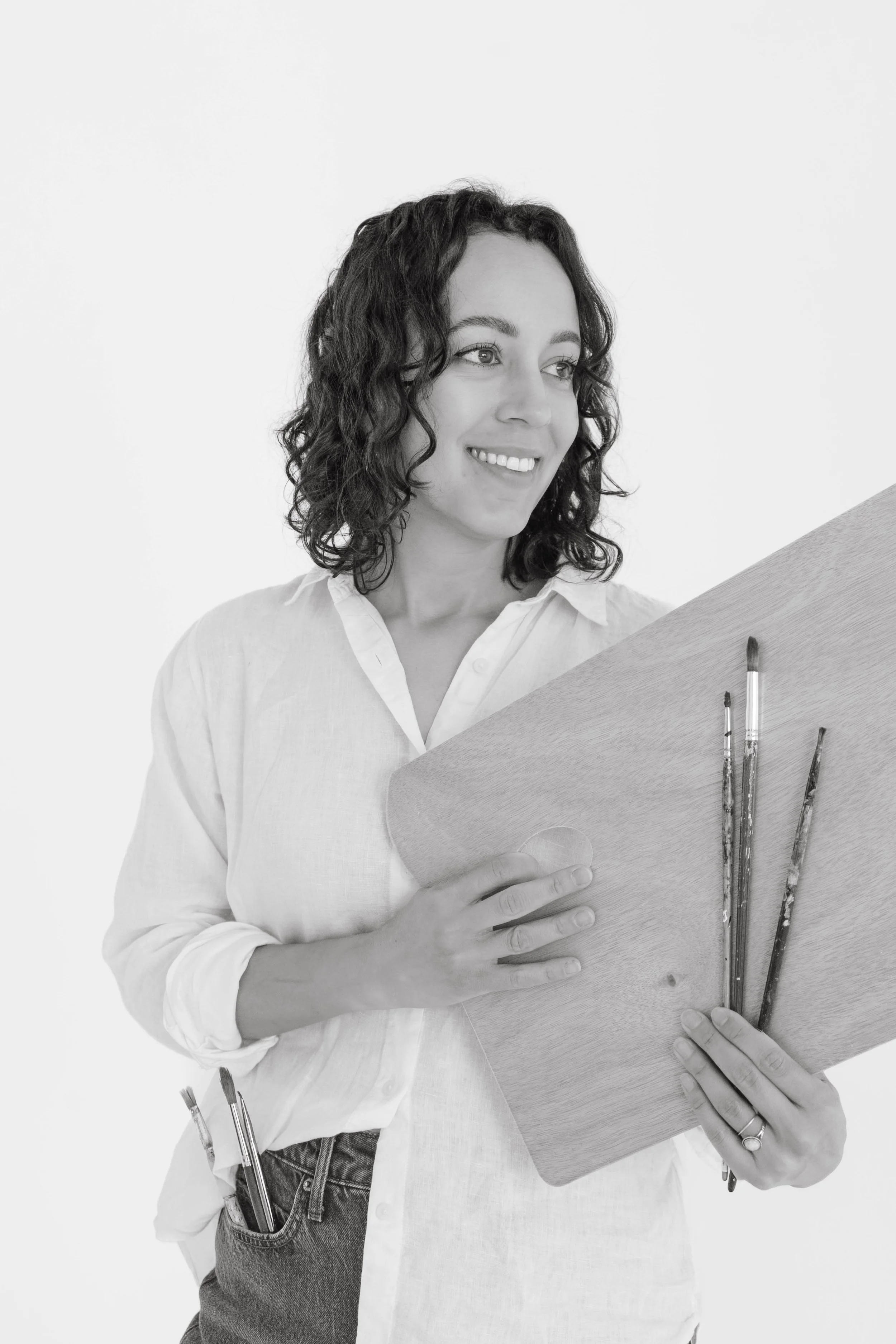 Black and white photo of a woman with curly hair smiling while holding a sketchbook. She is wearing a light-colored button-up shirt and has pens in a pocket on her jeans.