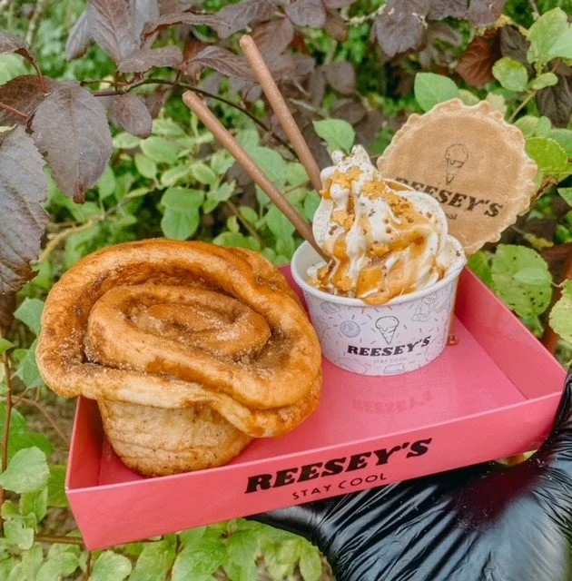 Cinnamon roll and vanilla ice cream with caramel sauce, whipped cream, and waffle cone piece in a Reese's branded cup on a pink tray, outdoors among greenery.