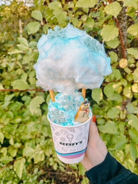 Hand holding a cup with a snowman decoration, topped with cotton candy, against a background of green leaves.