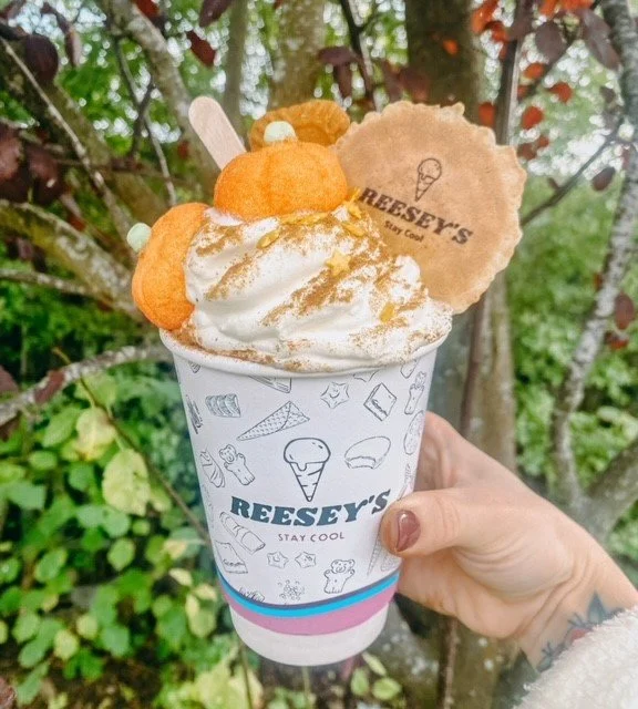 Hand holding a Reese's branded ice cream cup topped with whipped cream, a Reese's cookie, a pumpkin-shaped sweet treat, and a pumpkin lollipop, outdoors near trees.