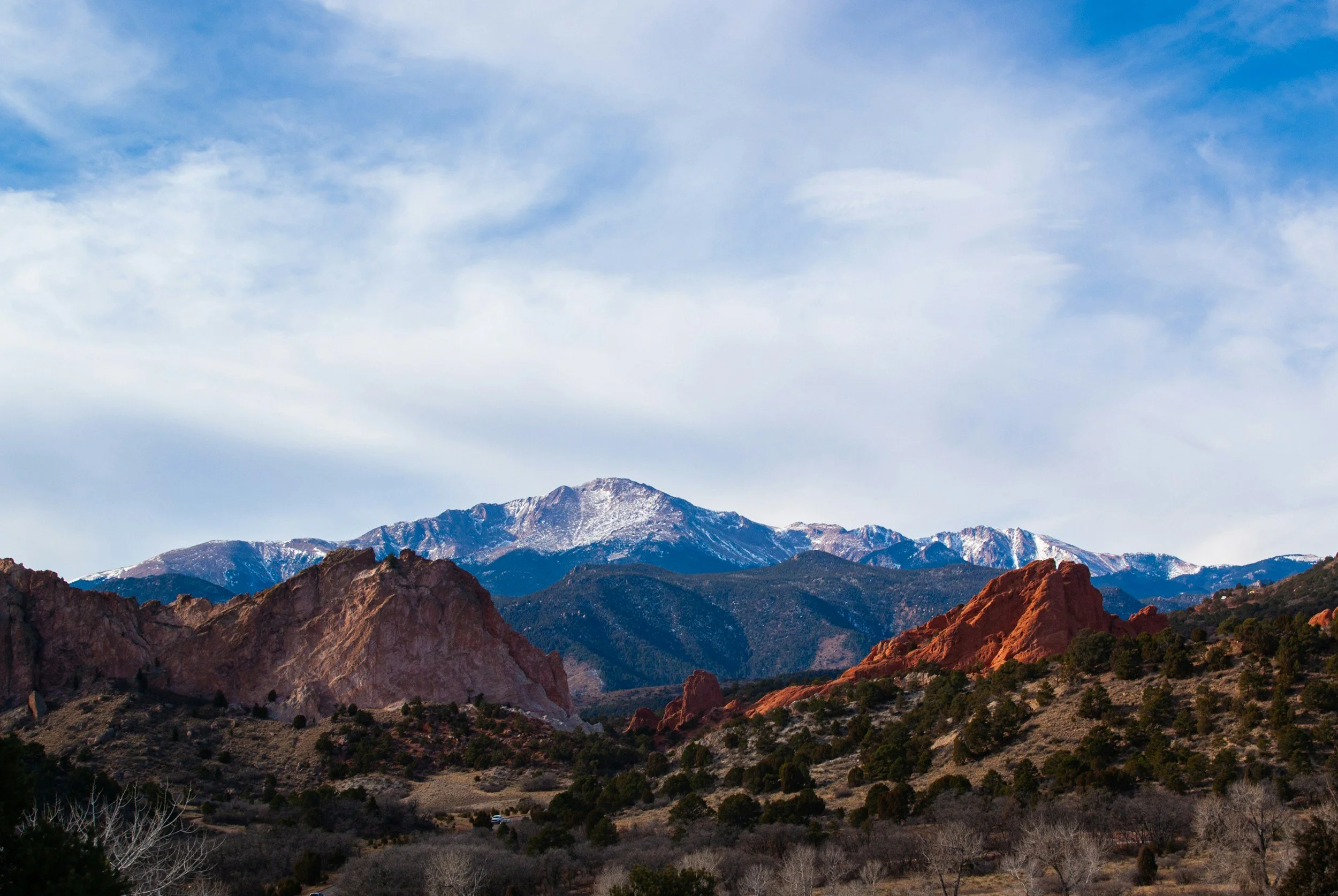 Mountain range with snow-capped peaks under a blue sky with few clouds, reddish rocks and sparse trees in the foreground.
