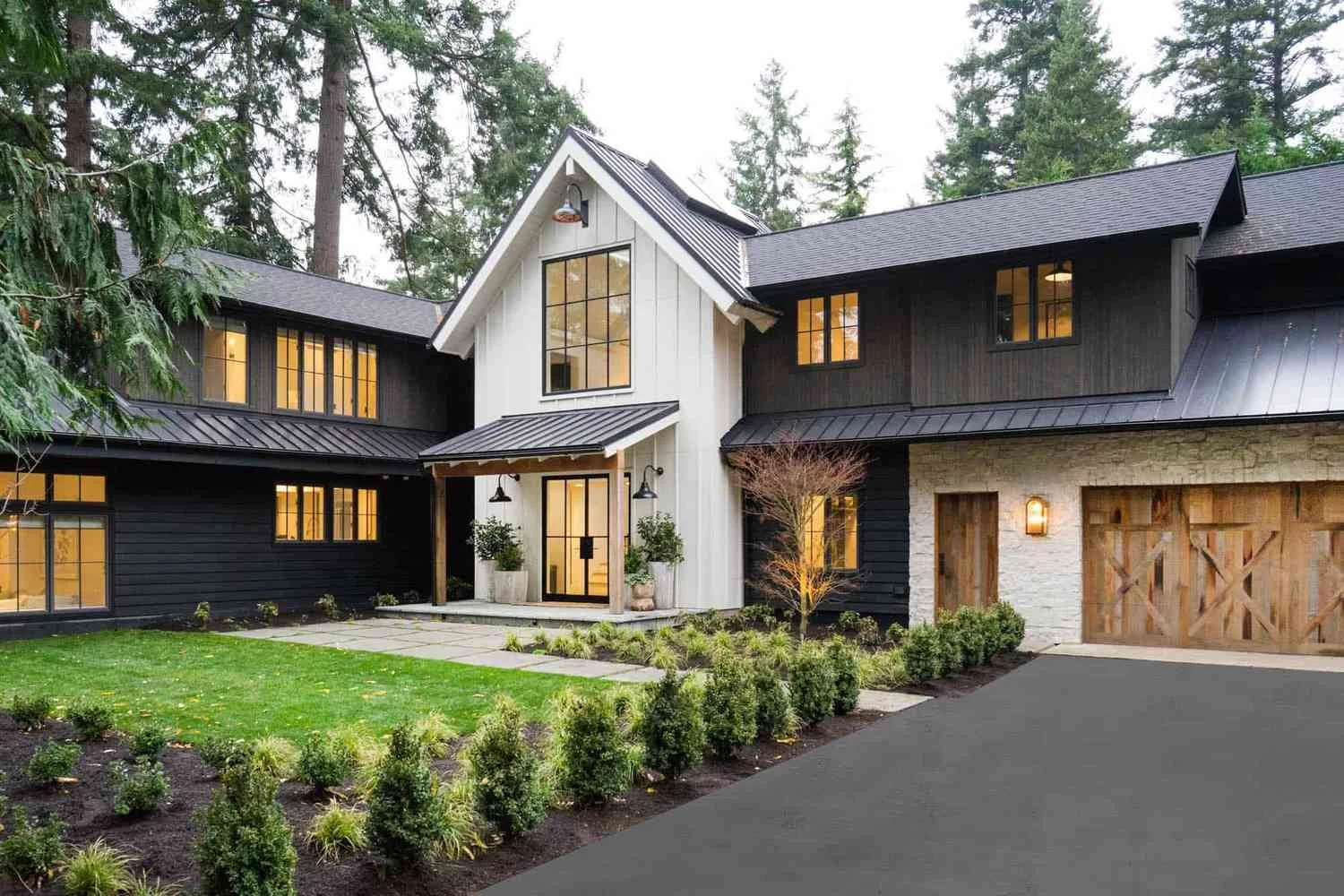 Modern black and white house with a metal roof, large windows, and a wooden garage door, surrounded by a landscaped garden with shrubs and a lawn.