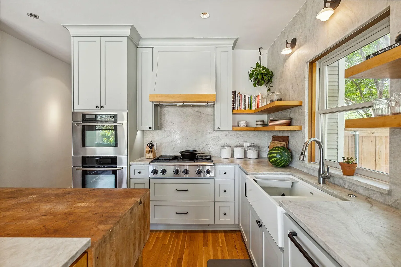 A kitchen with white cabinets, a marble backsplash, open wooden shelves, a window with a view of greenery, and various kitchen appliances and decor.