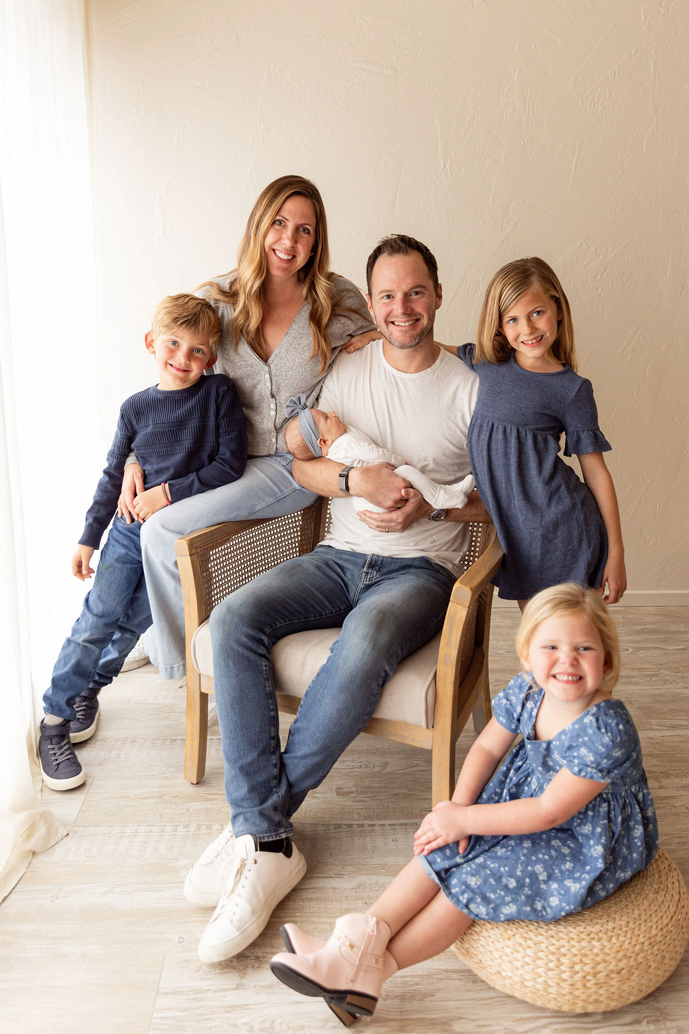 A smiling family of seven, including two adults, two young girls, two young boys, and a newborn, posing together in a bright room with a beige wall and light-colored wooden floor.