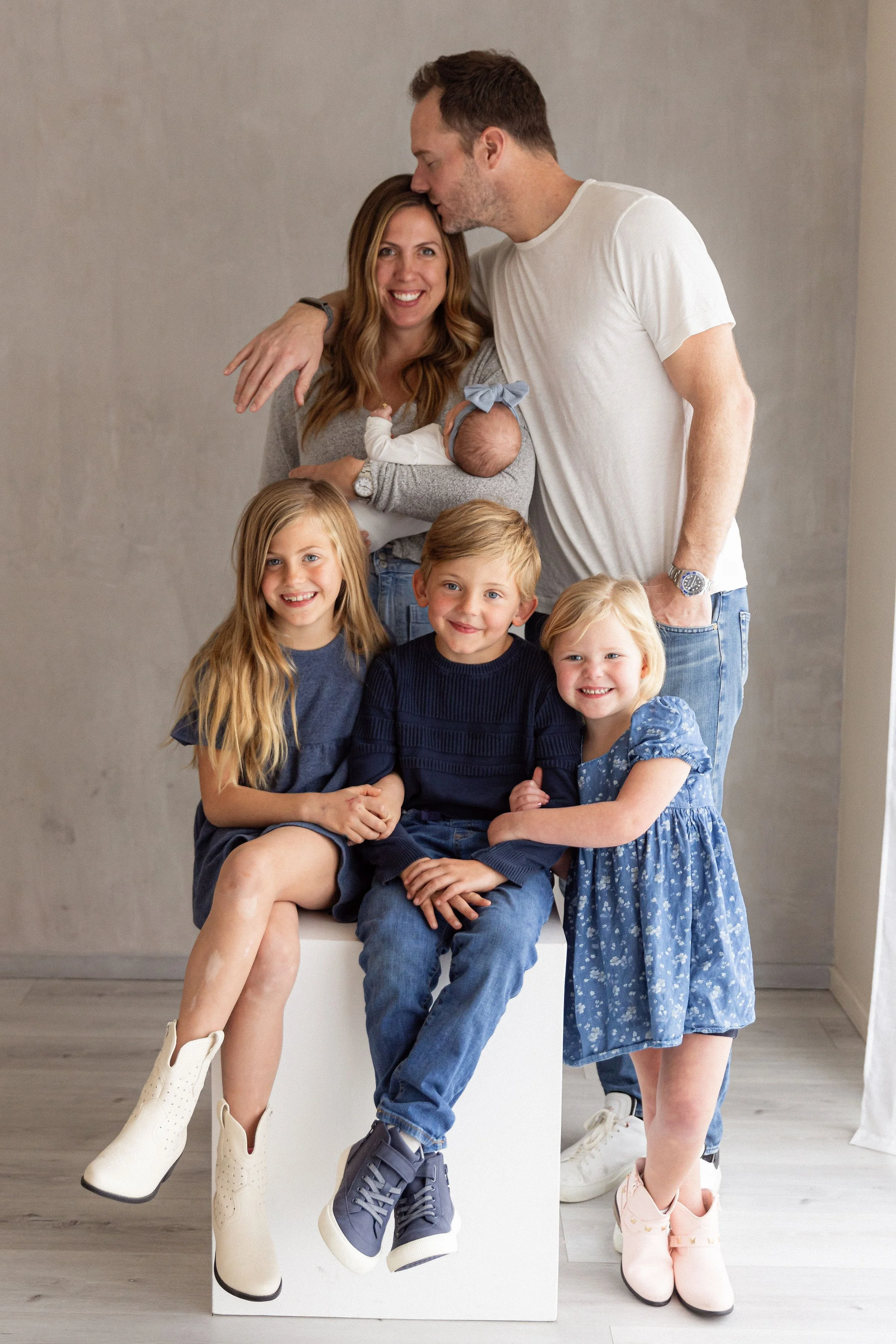 A family of six posing together. The mother holds a newborn baby, and the father is kissing her on the forehead. Three children are seated in front, smiling. The second child from the left is sitting on the father's lap, and a young girl stands beside the mother, smiling. The family is dressed casually, and they are indoors against a plain background.