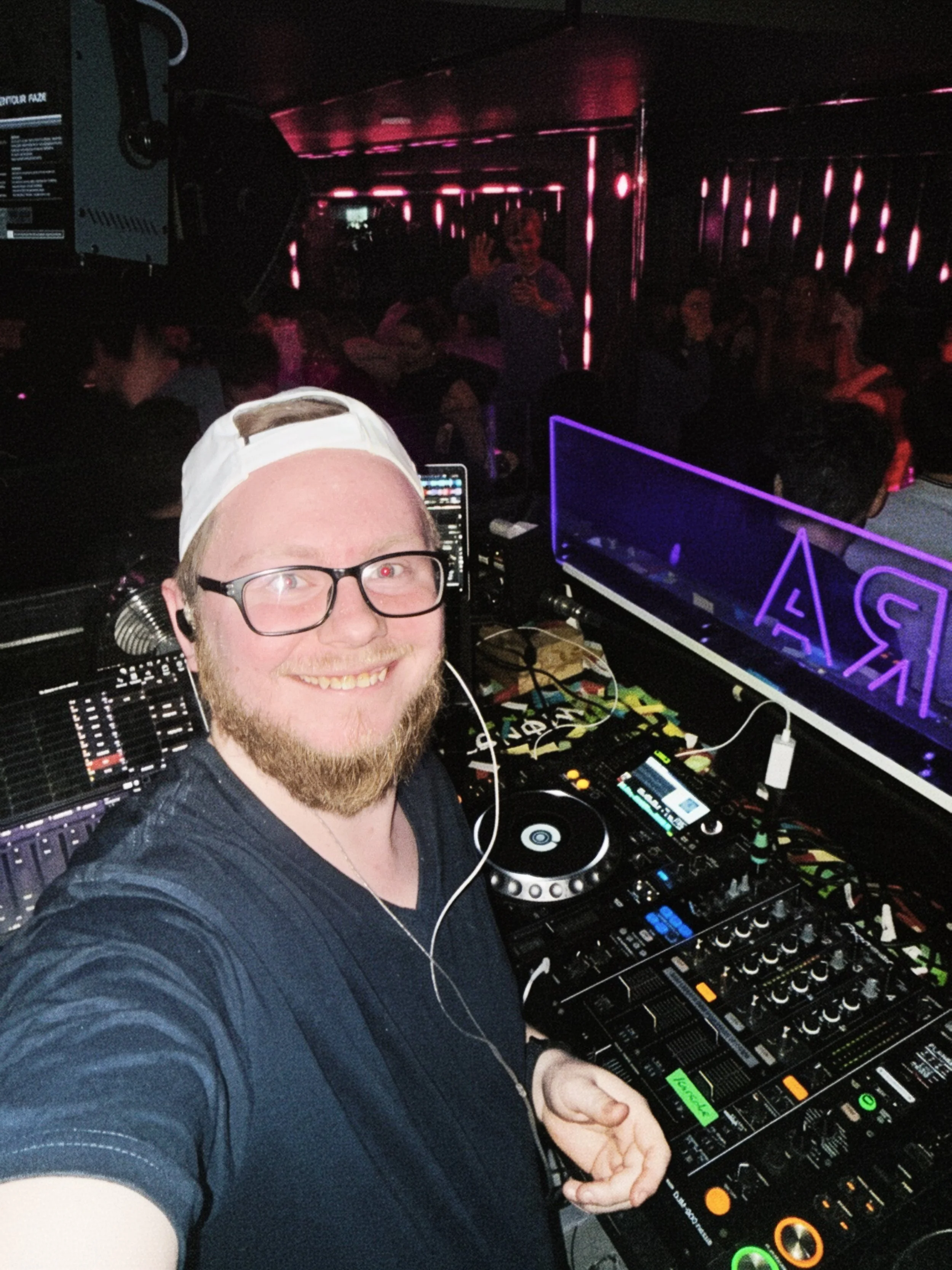 A man with glasses and a white cap smiling at a DJ setup, holding a pair of headphones, in a dimly lit club with people in the background and neon lighting.