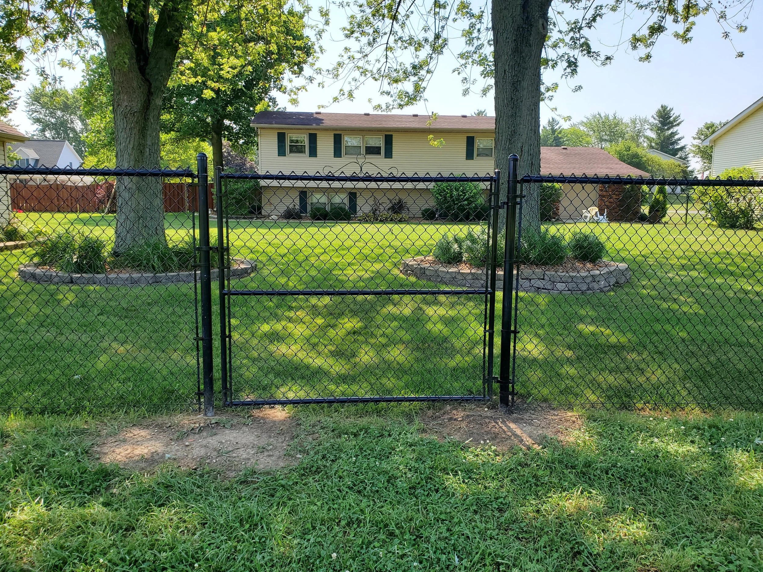 A black metal fence with a gate in front of a two-story house with beige siding and dark shutters, surrounded by green grass, trees, and bushes on a sunny day.