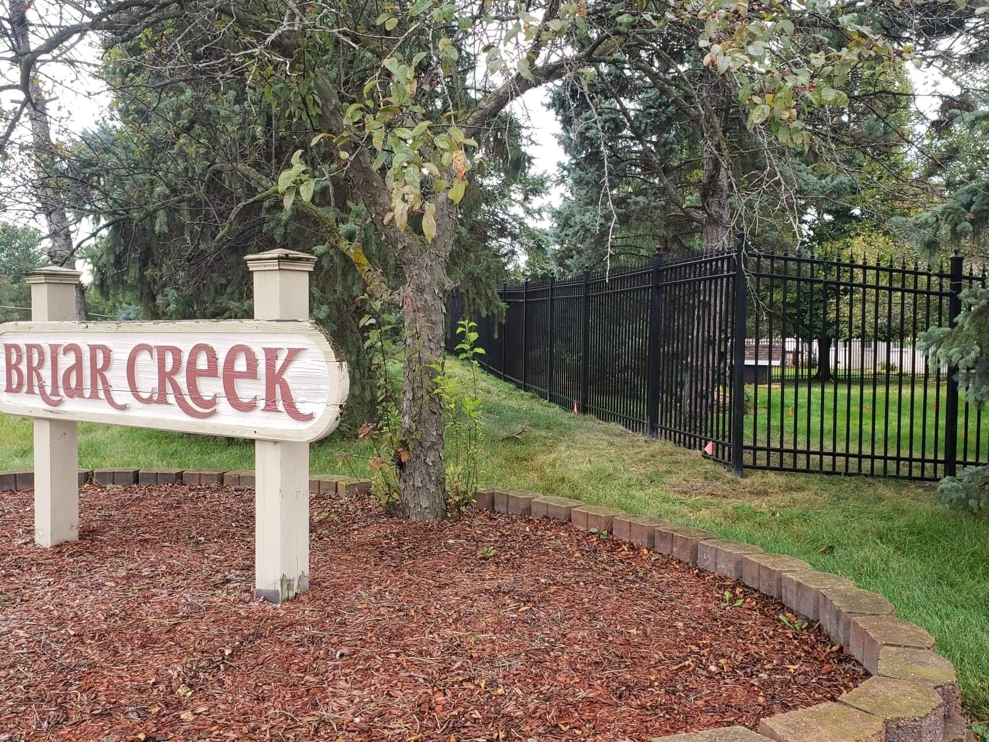 A park scene featuring a wooden sign reading 'Briar Creek,' a tree with green leaves, a black metal fence, and a grassy area with trees in the background.