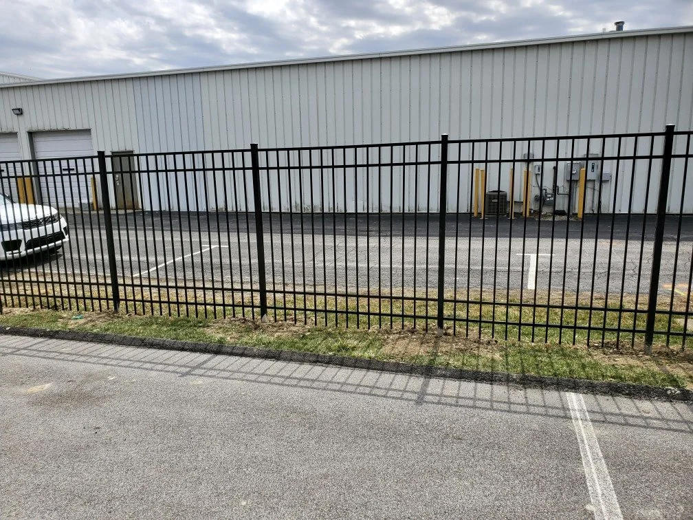 An empty parking lot with a black metal fence separating it from the sidewalk, a white van, and a large industrial building with garage doors in the background.
