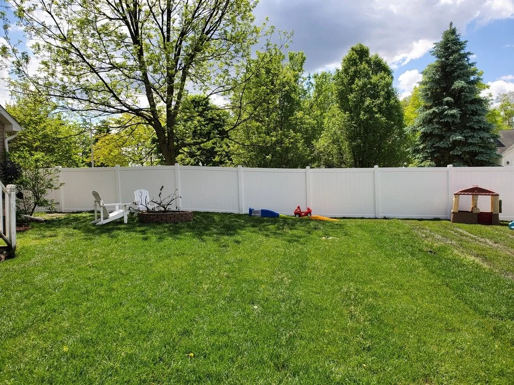 A backyard with a white fence, lush green grass, and several trees, including a large one with sparse leaves. There are children's toys on the lawn, including a small plastic playhouse and a toy car with a slide attached. A white plastic chair is pla