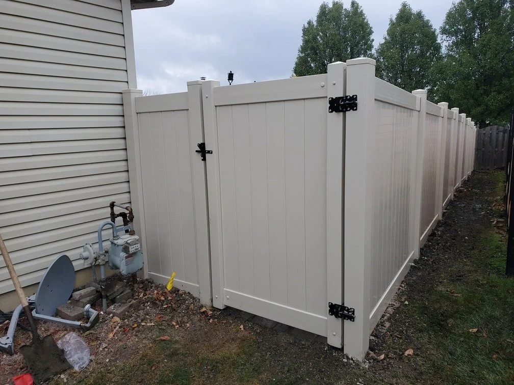 A white vinyl privacy fence surrounding a backyard with trees in the background. A corner of the house with beige siding and a gas meter with pipes are visible on the left side.