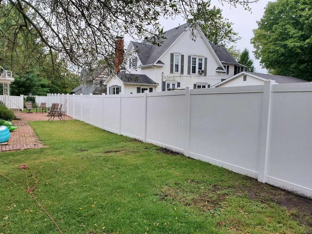 A backyard with a white privacy fence, green grass, outdoor seating, and a large white house with gray roof in the background.